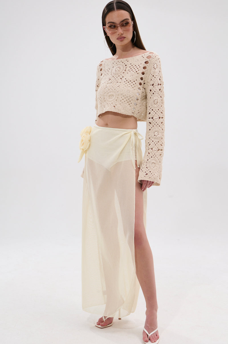 A woman in sunglasses, a cream crochet crop top, and the CROCHET BEAUTY COVER UP SKIRT—a sheer pale yellow wrap with floral detail—stands against a white background in white heels, ready for any beach-to-brunch moment. 6