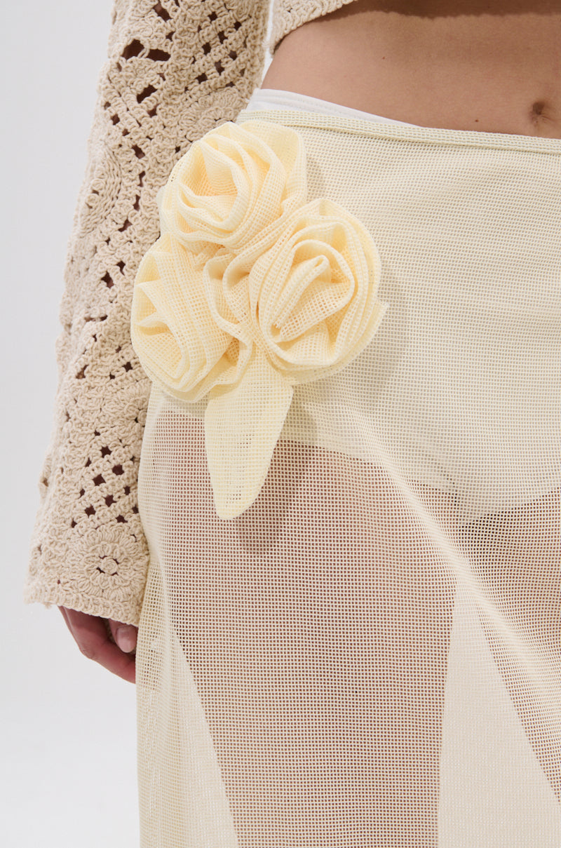 A close-up shows someone in a cream crochet sleeve and the CROCHET BEAUTY COVER UP SKIRT—a sheer mesh wrap skirt featuring large pale yellow flowers at the waist. Ideal for an effortlessly chic look on a plain, light backdrop. 7
