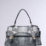 The TOO CROC TO HANDLE PURSE showcases a chic design with metallic faux crocodile leather, adorned with decorative buckles on the front. Its structured rectangular shape and sleek, modern aesthetic are complemented by a short handle and detachable shoulder strap, all set against a plain light gray background.