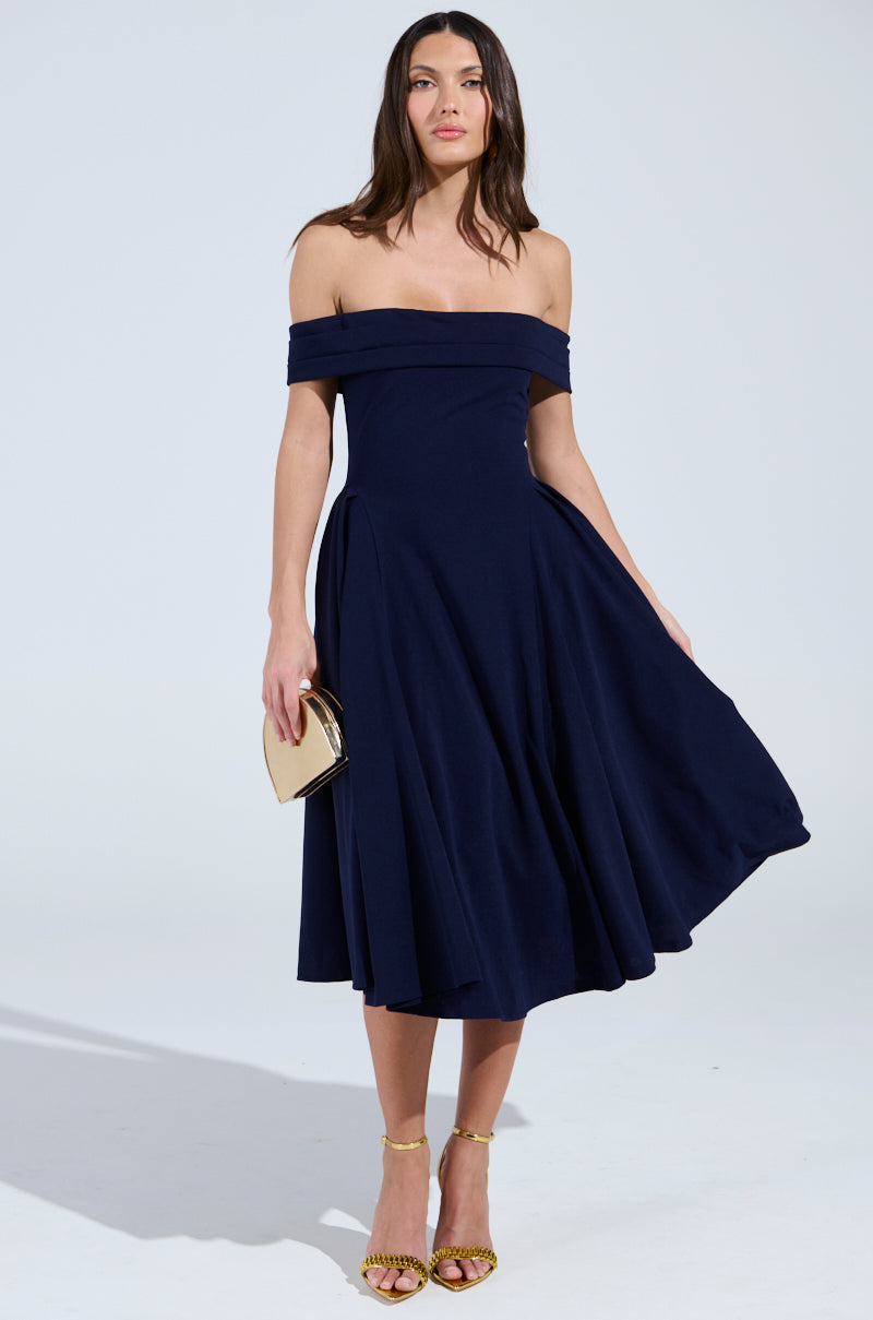 A woman in an off-the-shoulder ELEVATE ME PLEATED MIDI DRESS stands against a light gray background, holding a small gold clutch. The navy blue dress flares slightly for a flowing look, and she pairs it with gold strappy heels.