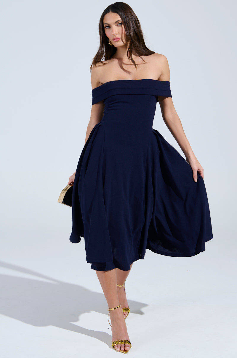 A woman confidently poses in the ELEVATE ME PLEATED MIDI DRESS, a navy blue fit 'n flare with an off-the-shoulder style. She holds a small gold clutch in her right hand, standing against a plain background and wearing yellow strappy heels.