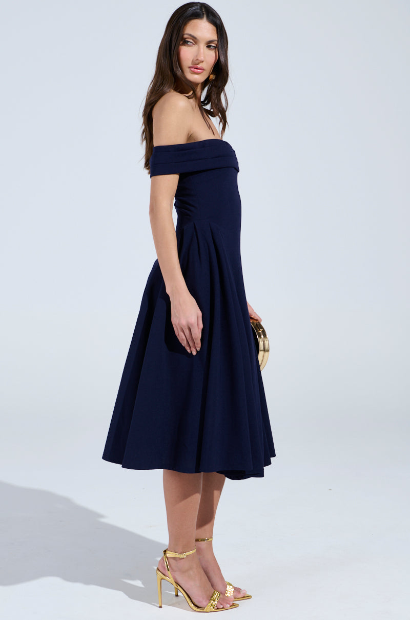 A woman in an off-the-shoulder navy blue ELEVATE ME PLEATED MIDI DRESS and gold high heels holds a small round clutch, posing sideways against a light background with her long hair cascading down her back.