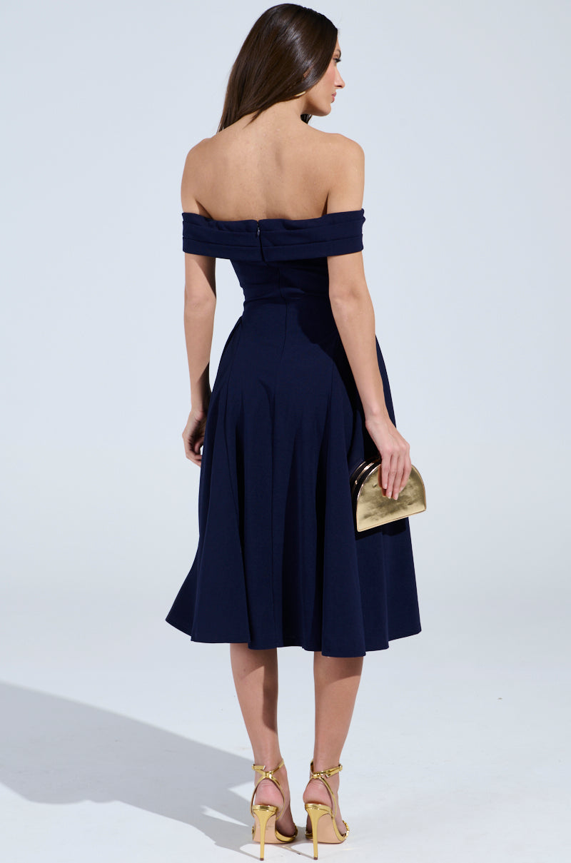A woman with long brown hair is seen from the back, wearing the ELEVATE ME PLEATED MIDI DRESS in navy blue and gold high-heeled sandals, holding a small gold clutch in her left hand against a plain white background.