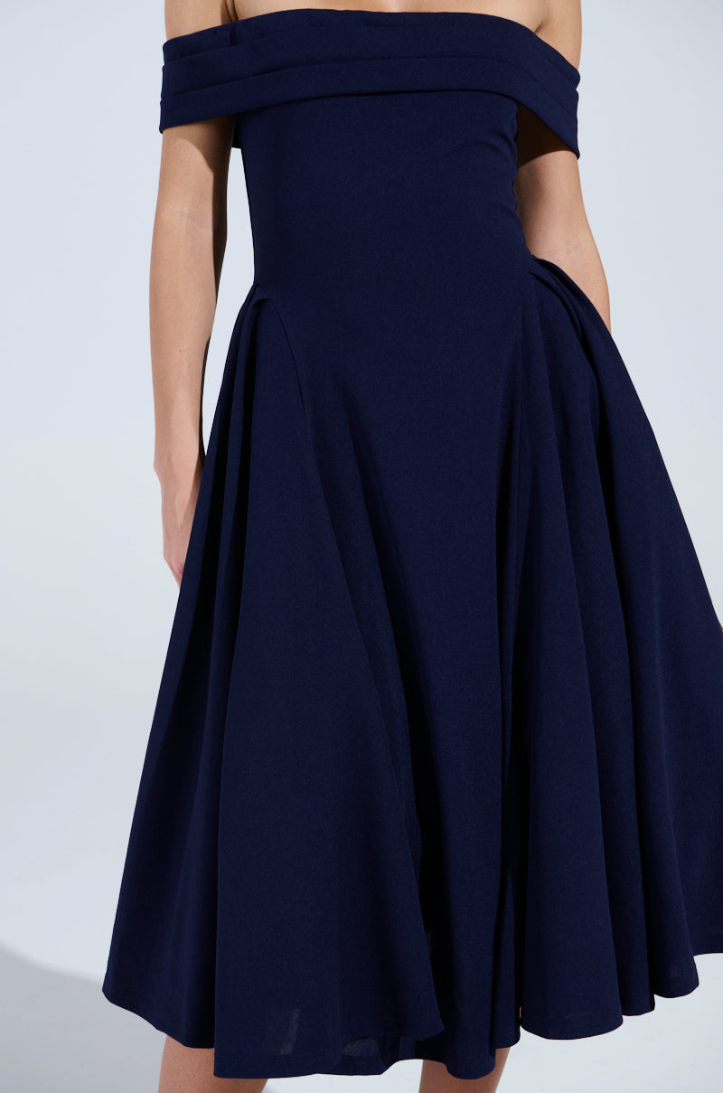 The ELEVATE ME PLEATED MIDI DRESS in dark blue showcases its elegant off-the-shoulder fit and flared pleated midi skirt, capturing the spotlight against a plain backdrop while keeping the person's face unseen.