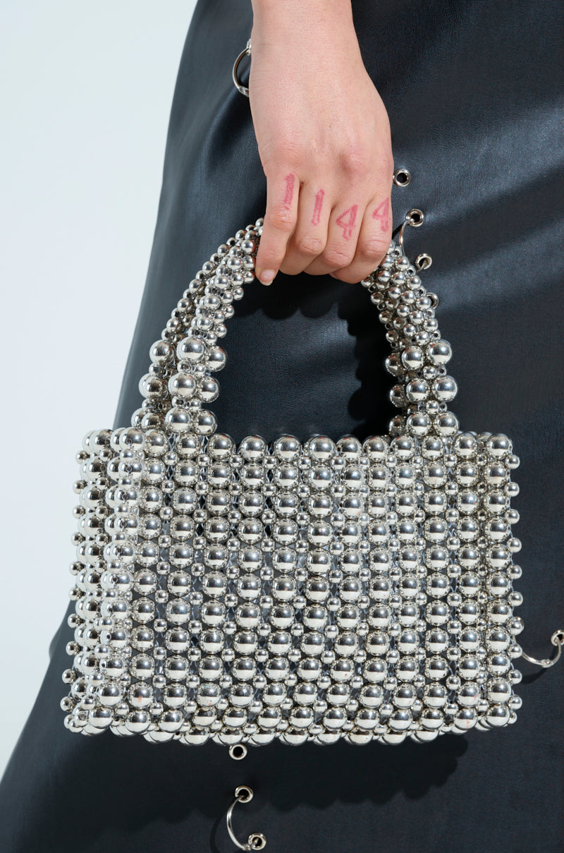 A person carries the IM SO IT GIRL BEADED PURSE, crafted from metallic silver beads with dual beaded handles, while donning a black skirt accented with small silver rings and their fingers embellished with letters and symbols.