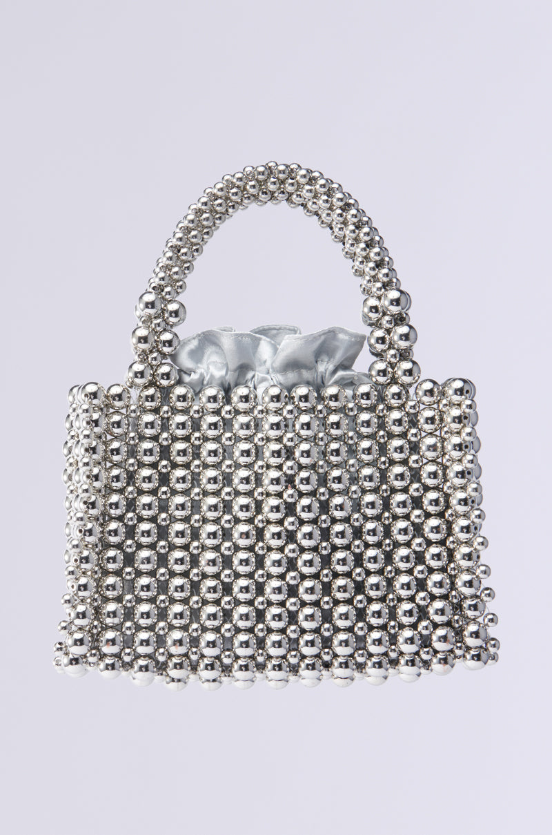 The IM SO IT GIRL BEADED PURSE, crafted from interlinked metallic silver beads, features a bold design with a short handle and shiny silver lining. Its plain light gray background ensures the spotlight stays on this standout piece.