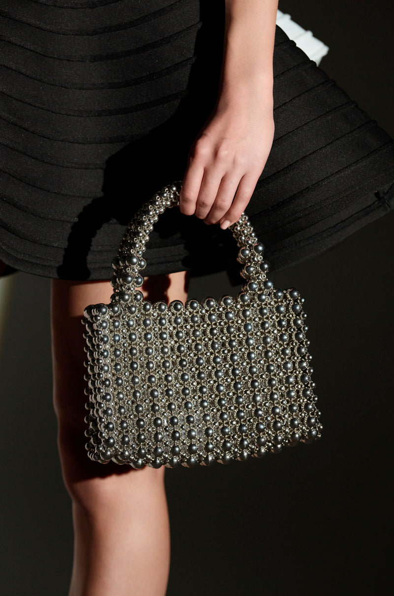 A person holds the IM SO IT GIRL BEADED PURSE, crafted with interconnected metallic silver beads. This classic hot-girl handbag features a short handle and contrasts elegantly against black, textured pleated fabric, with lighting beautifully highlighting its reflective surface.