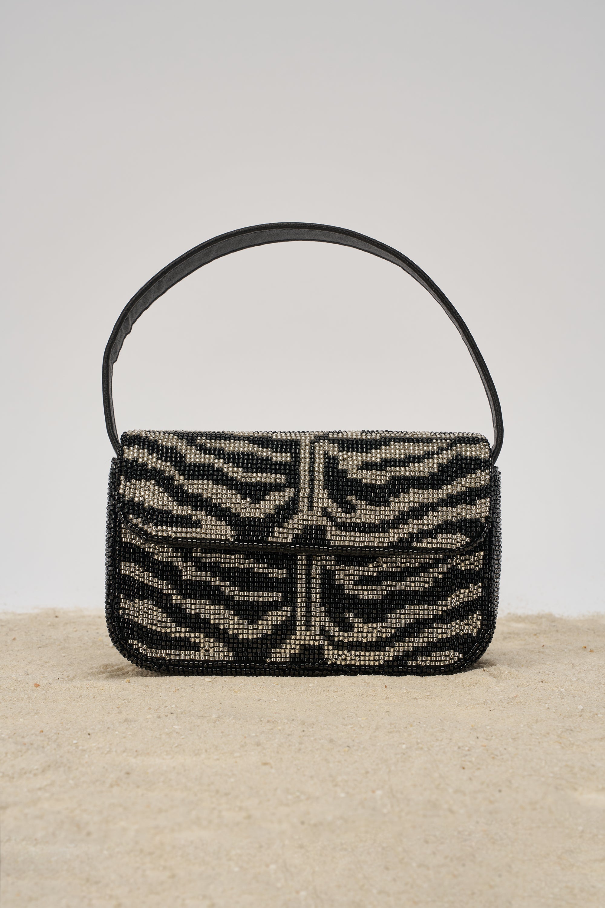 The VIVIANNE BEADED PURSE is a black and silver zebra-striped clutch with a short black strap, shown upright on sandy ground against a neutral background. 1