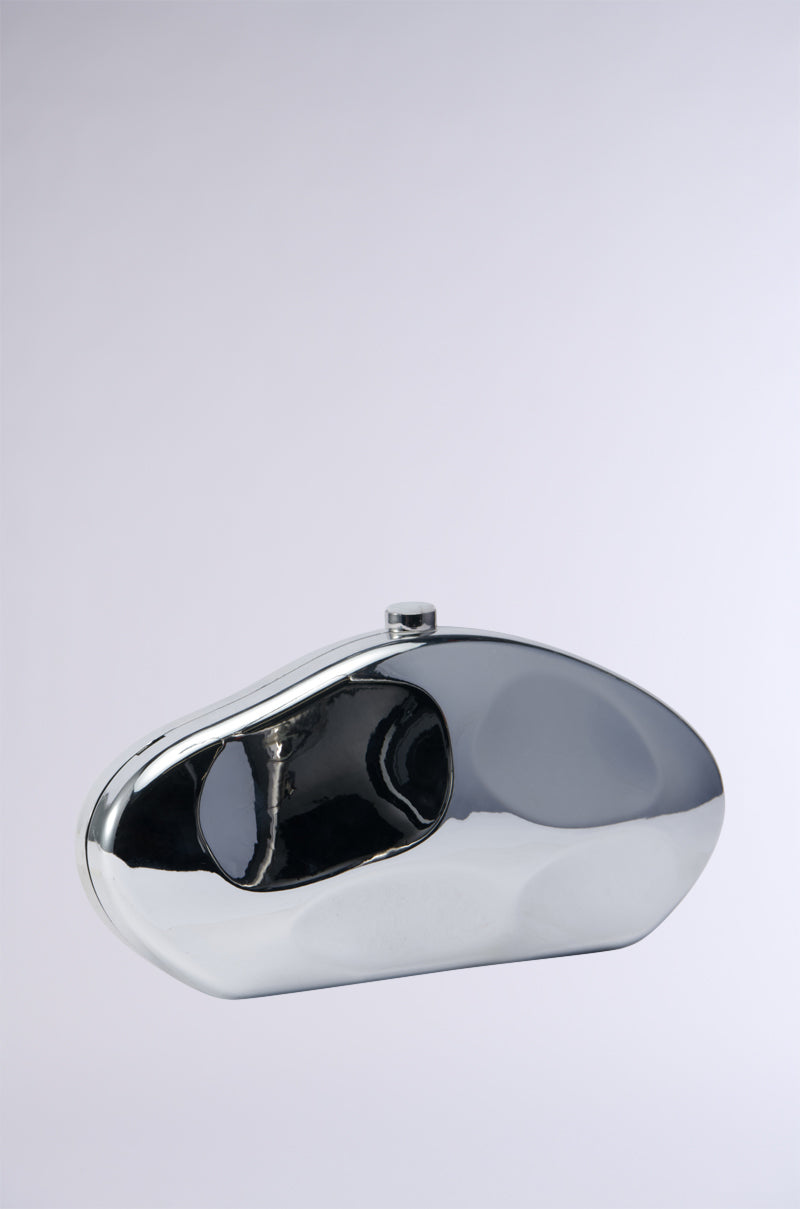 The BENT OUTTA SHAPE CLUTCH, a silver, asymmetrical clutch bag with a reflective, polished finish is displayed against a plain, light grey background. This metallic silver clutch features an abstract, modern design with smooth curves and a small clasp at the top. For added versatility, it comes with a detachable chain link strap. 0