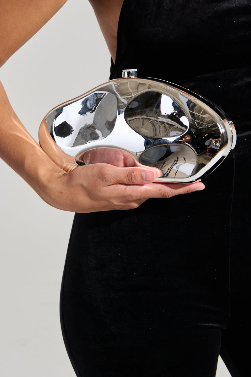 A person dressed in a black outfit holds the BENT OUTTA SHAPE CLUTCH, an abstract design bag featuring a metallic silver finish. This purse has an irregular, organic shape with reflective surfaces and includes a detachable chain link strap. The image is cropped to focus on the hand holding the clutch, captured from the neck to the knees. 1