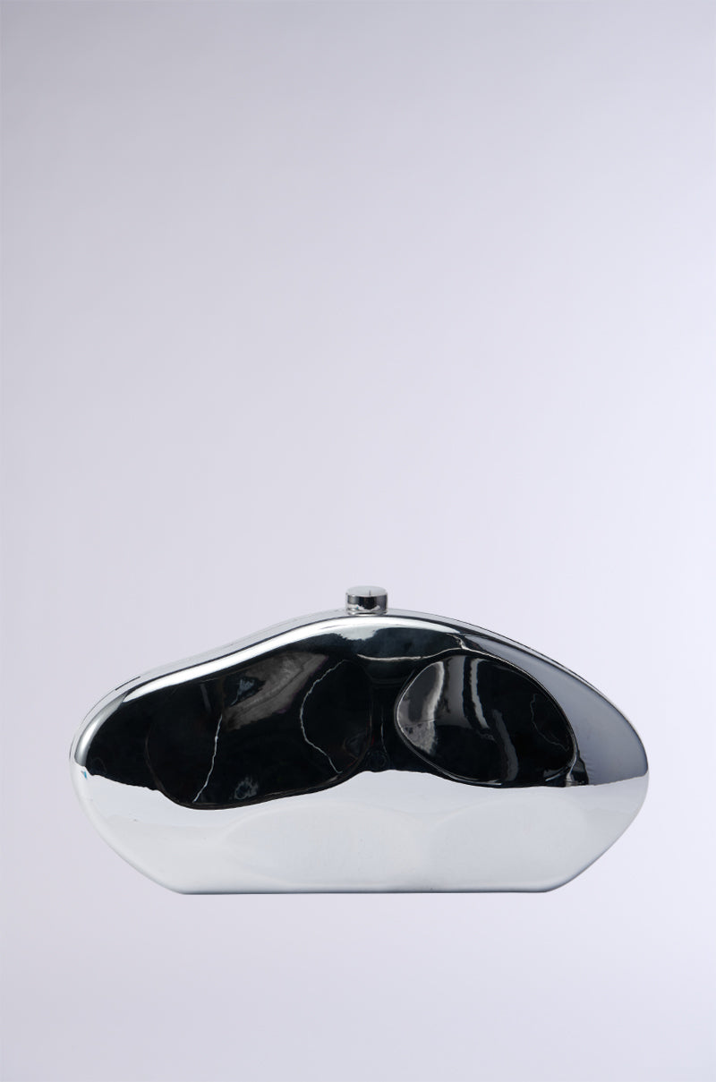 The BENT OUTTA SHAPE CLUTCH is a sleek, metallic silver clutch bag with an abstract shape, set against a light gray background. It boasts a glossy surface and a minimalist design, featuring a small clasp at the top for closure. For added versatility, it includes a detachable chain link strap. 2