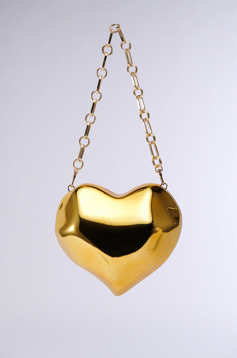 A glossy, gold-colored heart-shaped handbag with a coordinating gold chain strap. The HEART TOO BIG CHROME CLUTCH showcases a sleek and reflective surface, elegantly suspended against a simple white background.