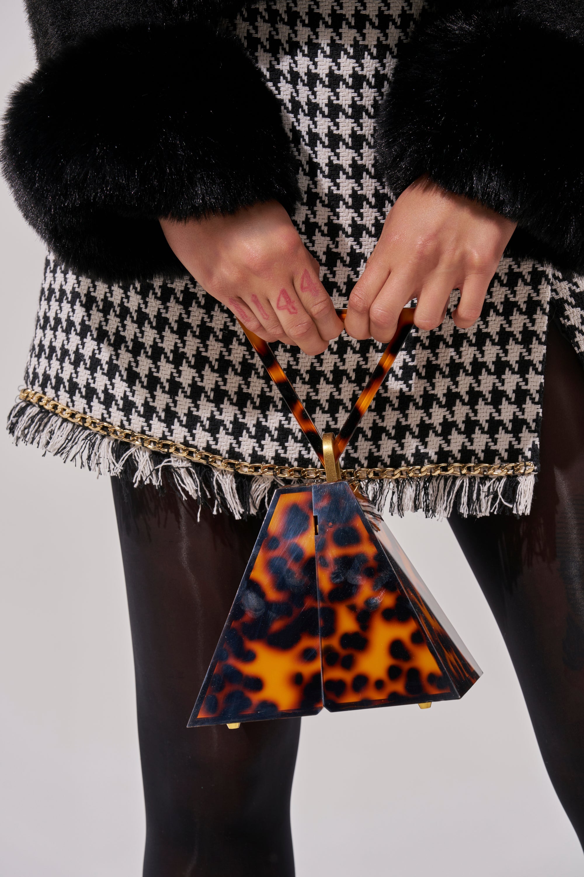 A person in a houndstooth skirt with black faux fur cuffs holds the TOP OF THE PYRAMID PURSE, a small triangular tortoiseshell handbag. They're also wearing black tights and have pink nail polish. 2
