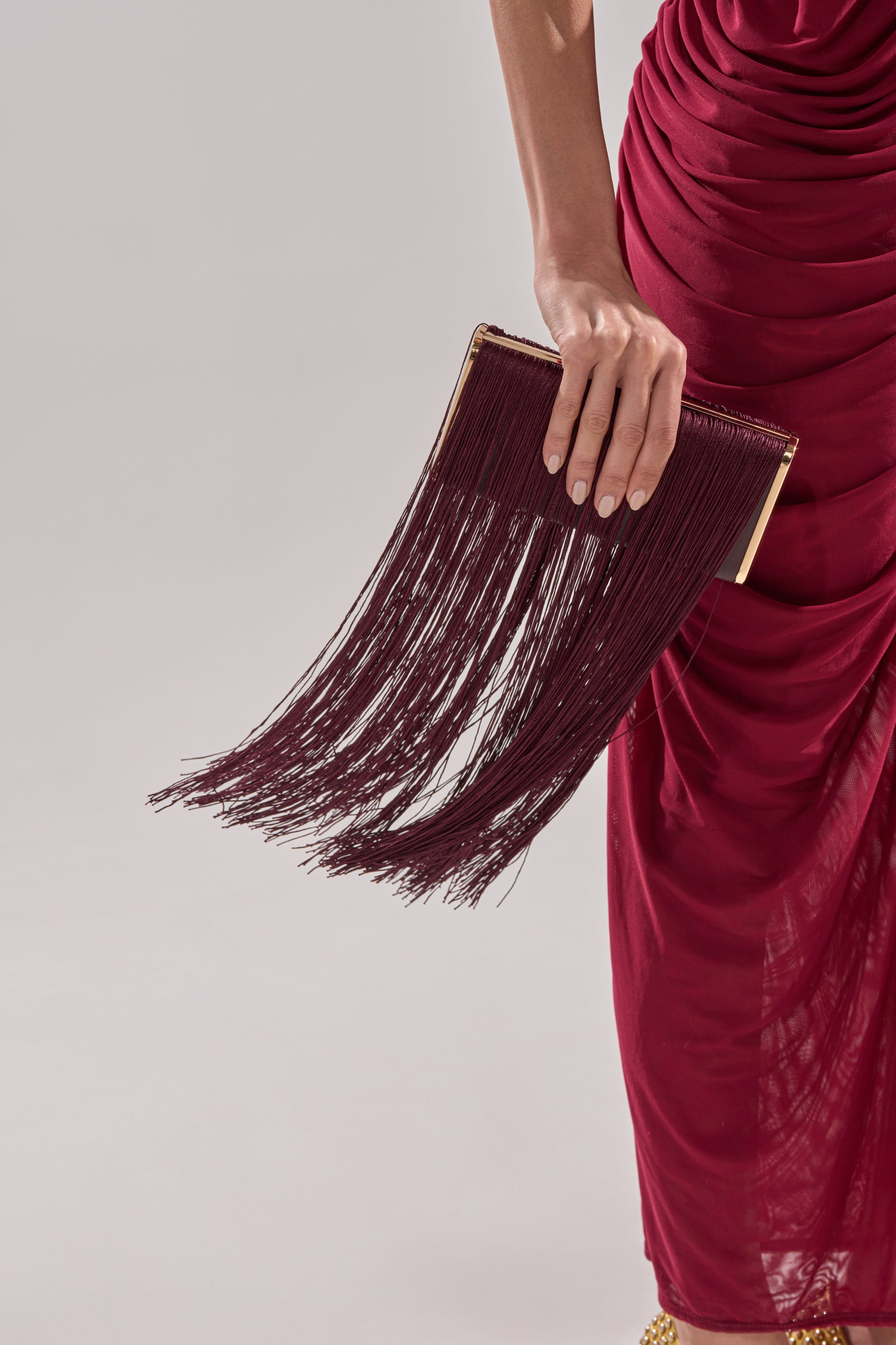 A person holds the IN THE FRINGE CLUTCH, a wine-colored bag with long fringe, while wearing a deep red ruched dress against a light gray background. 0