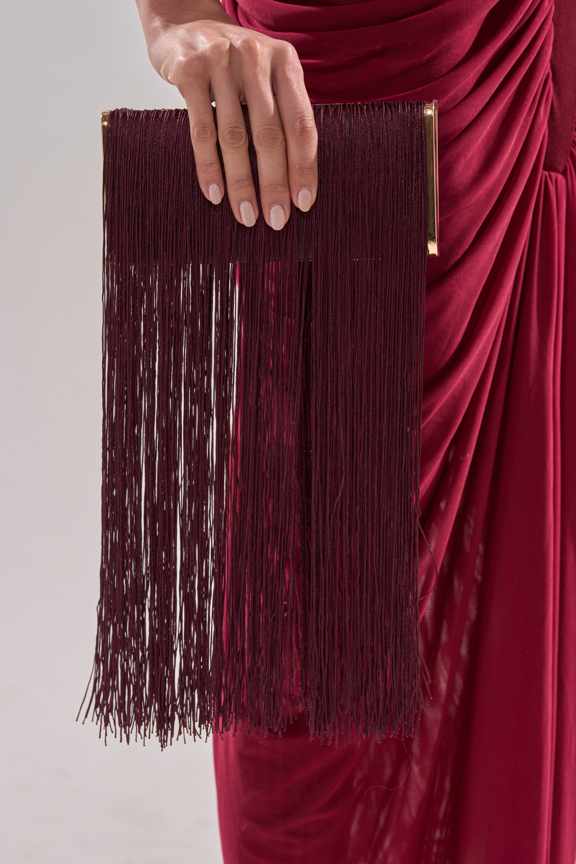 A person in a red, ruched dress holds the elegant IN THE FRINGE CLUTCH, featuring long hanging tassels and gold accents.