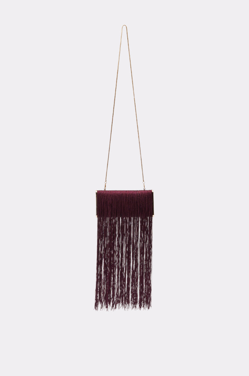 The IN THE FRINGE CLUTCH, in a rich wine shade with a long gold chain strap, hangs against a plain light gray background. 1