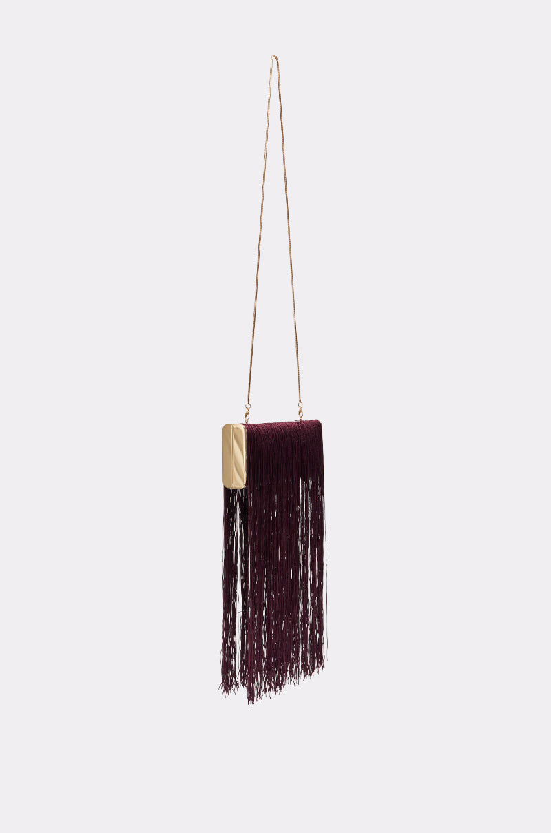 The IN THE FRINGE CLUTCH is a small rectangular evening bag with gold sides, a long gold chain strap, and dark burgundy fringe, shown against a plain light grey background.