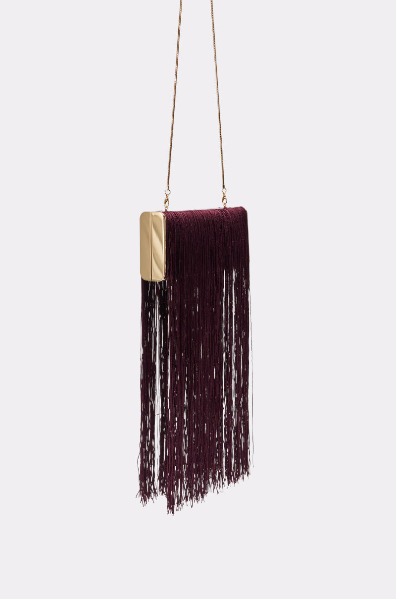 The IN THE FRINGE CLUTCH features a wine hue, gold rectangular frame, long burgundy fringe, and slim gold chain strap—an elegant evening bag that stands out against a plain light background.