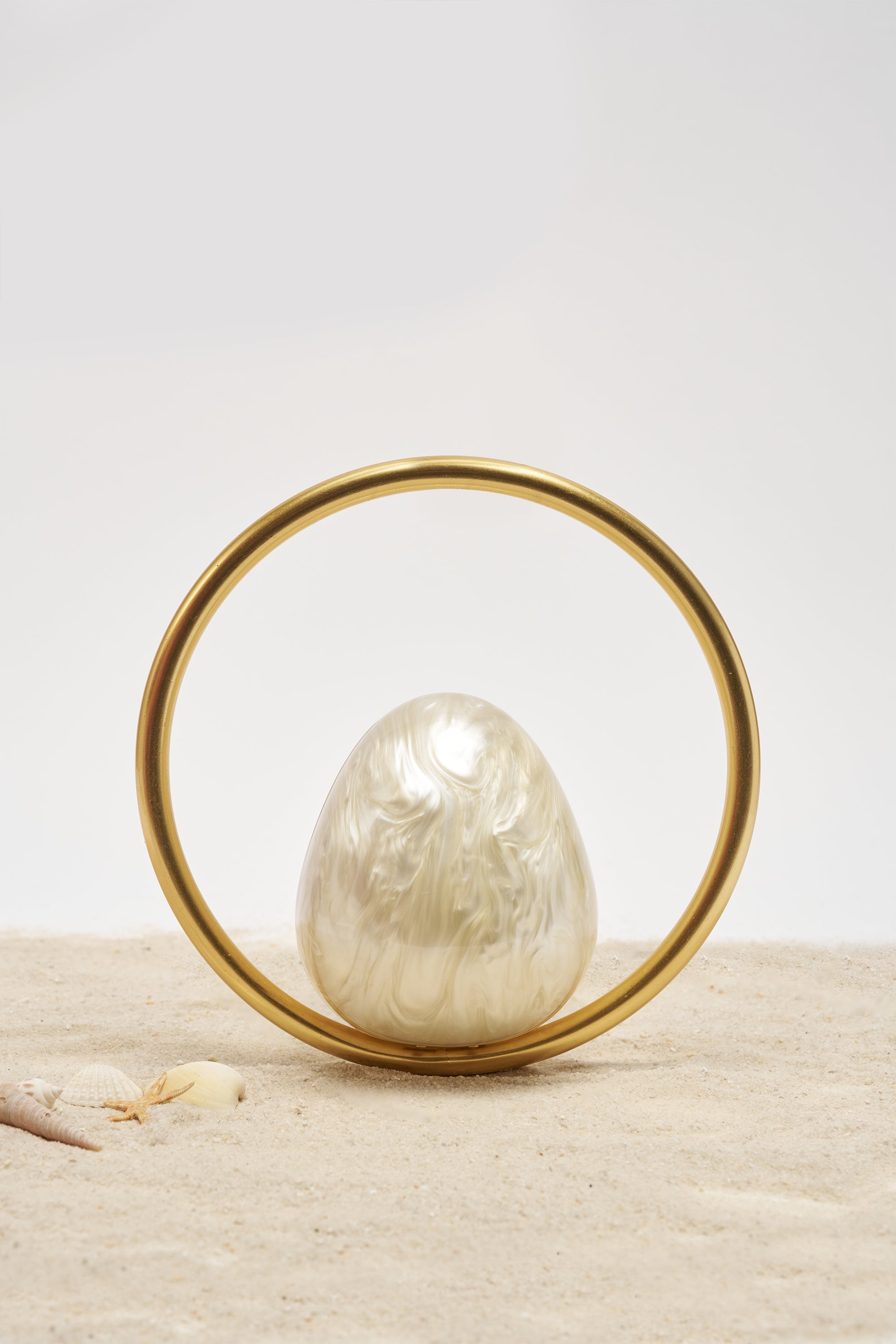 The ON MY MIND BAG, a pearlescent egg-shaped accessory with a gold-tone handle, rests on sand beside small seashells. The scene features a clean white background for a minimalistic look. 0