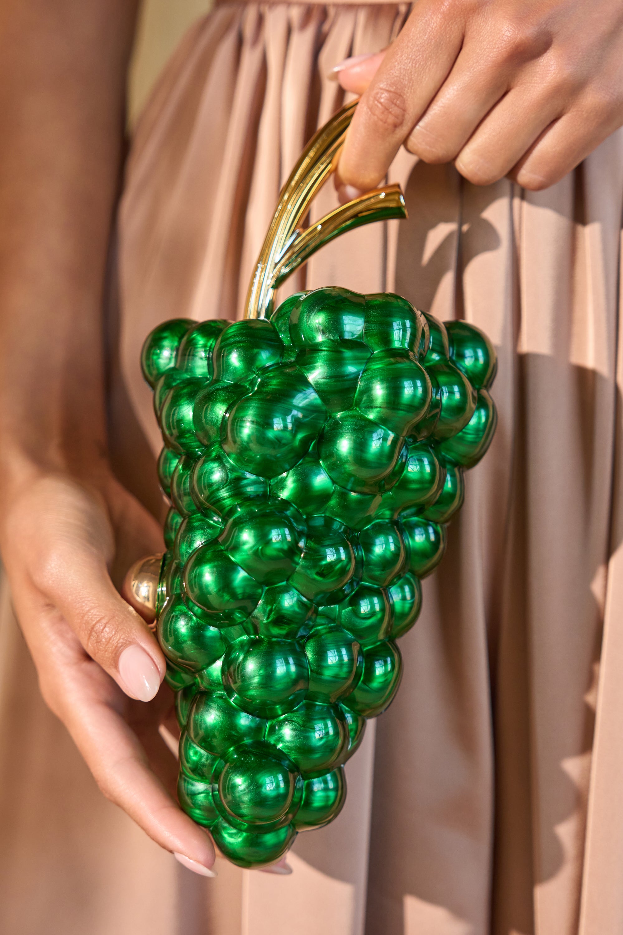 A person holds the I FEEL GRAPE CLUTCH—an acrylic bag with a gold stem handle—paired with a light beige pleated skirt or dress; it's an eye-catching party accessory for any occasion. 0