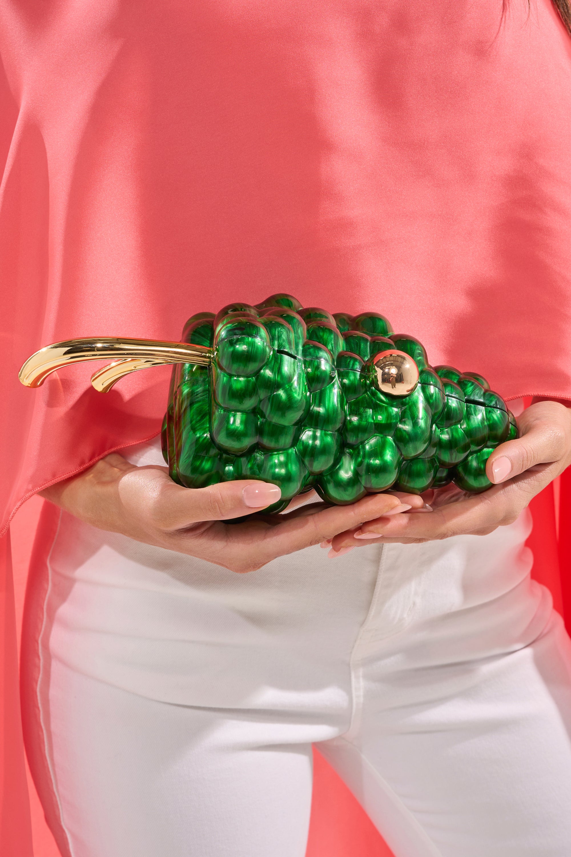 A person in a coral pink top and white pants holds the I FEEL GRAPE CLUTCH—a glossy acrylic bag with a gold stem, marbled finish, and a chic gold chain strap. 3
