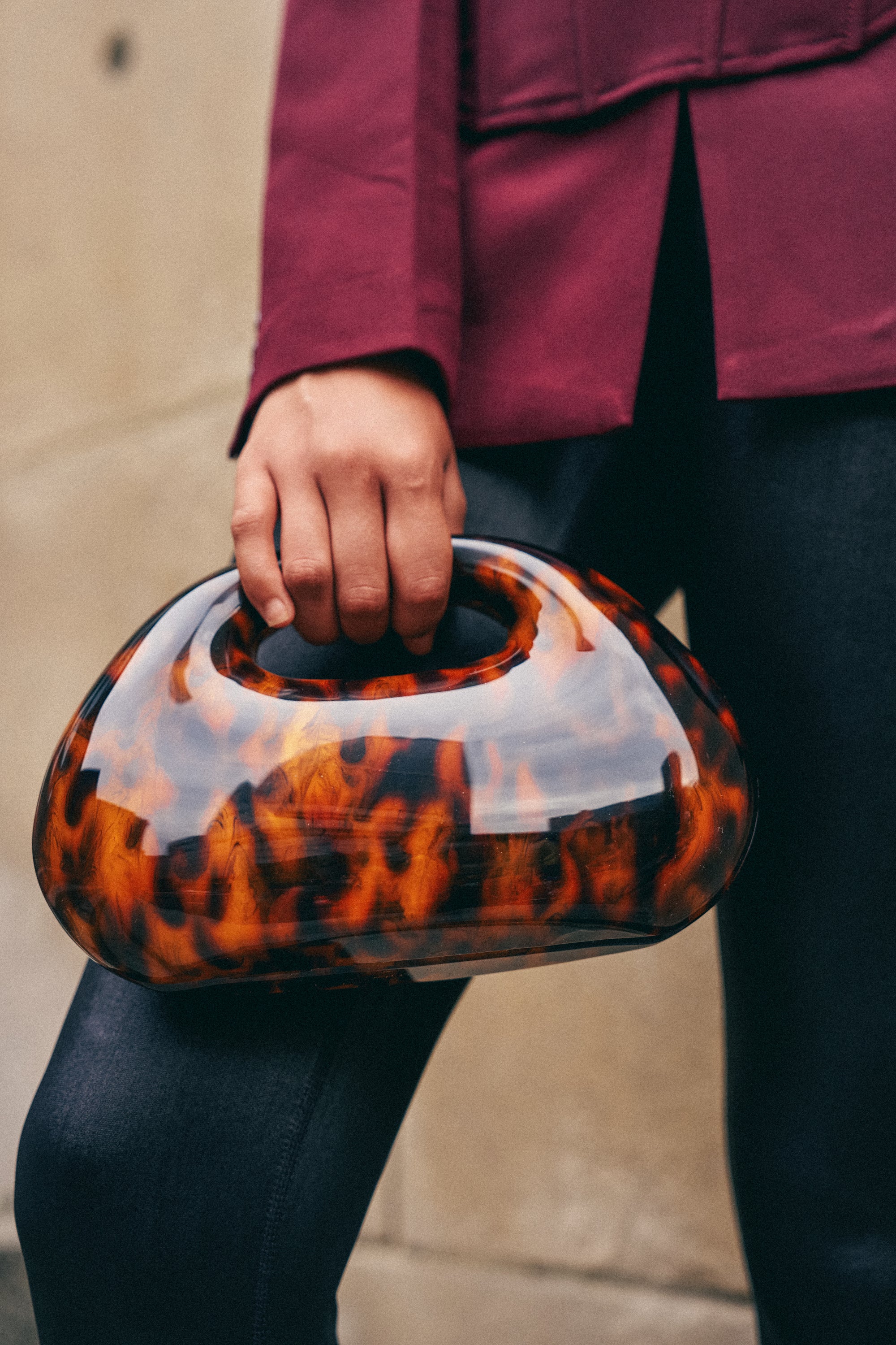 A person in a burgundy jacket and dark pants holds the TORTOISE CLUTCH, a glossy tortoiseshell acrylic handbag with a built-in handle. This retro-chic accessory features a detachable chain shoulder strap, effortlessly blending classic elegance with modern versatility.