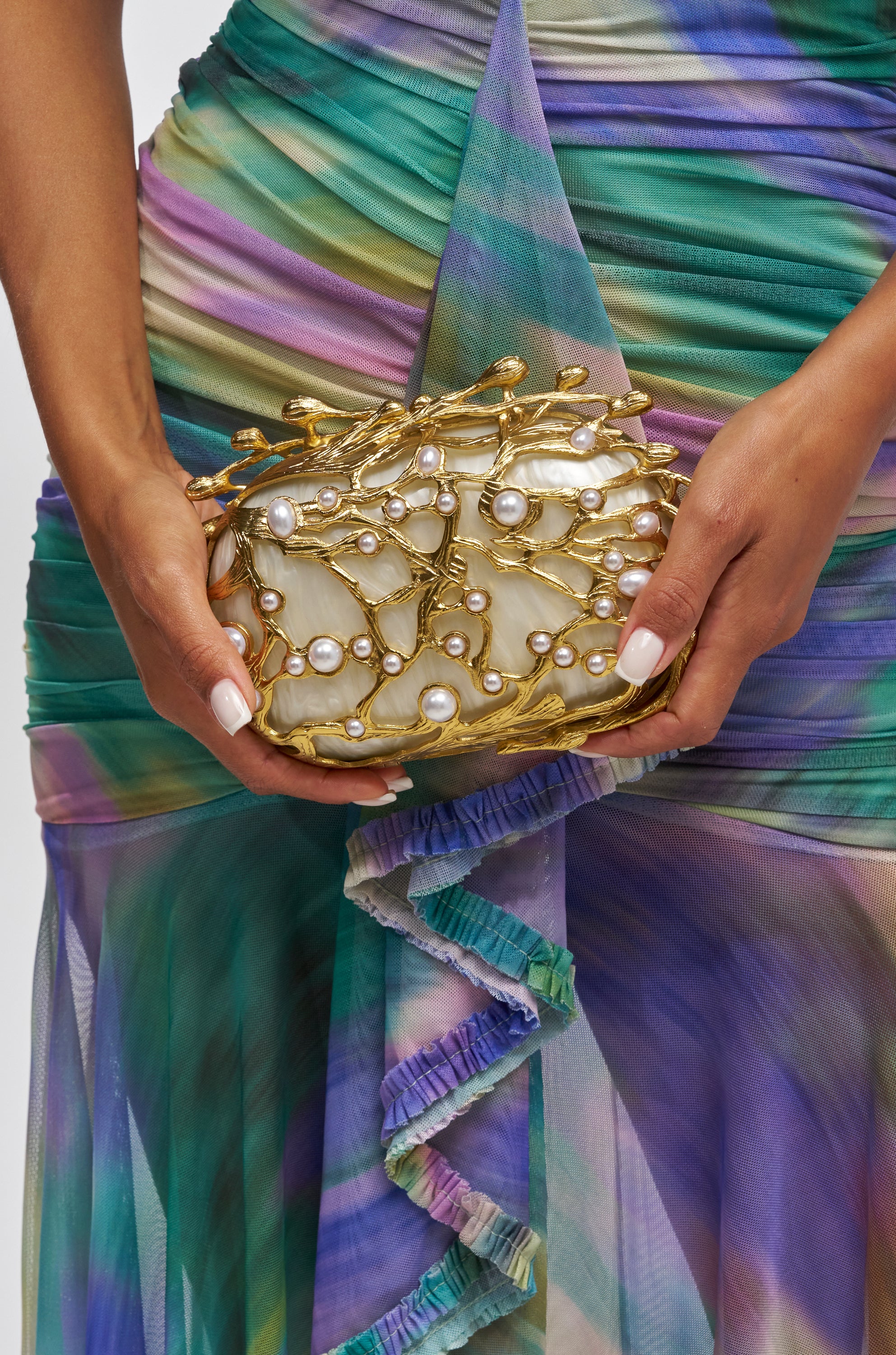 A person in a colorful, pleated dress holds the ALWAYS OUTSIDE CLUTCH—a gold branch clutch embellished with pearls. Their pale manicured nails complete the elegant look.