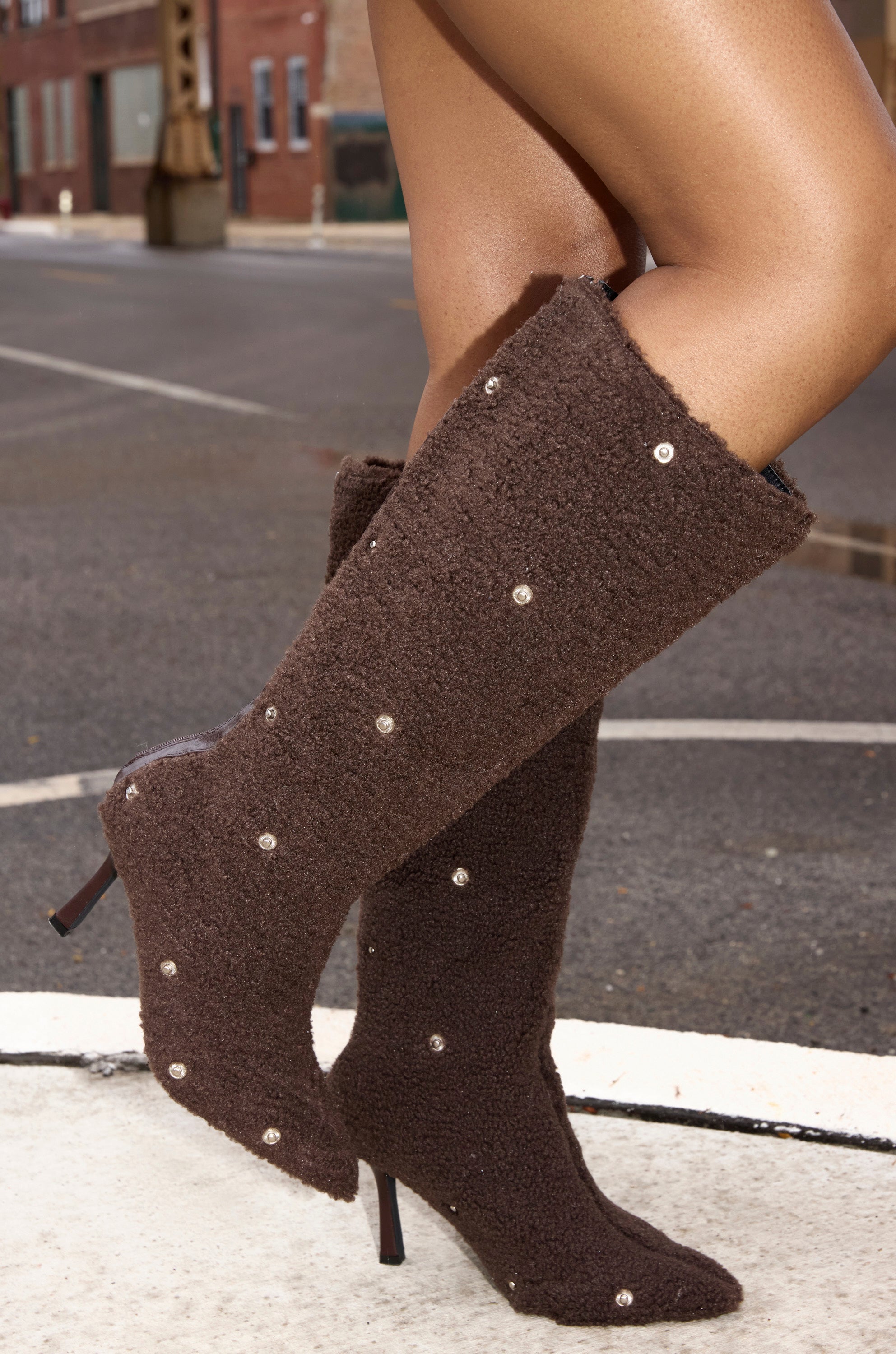 A close-up of lower legs and feet on a city sidewalk wearing the AZALEA WANG FABIAN BROWN SHERPA FOLDED DETAIL BOOT—knee-high, textured brown fabric, small studs, stiletto heel.