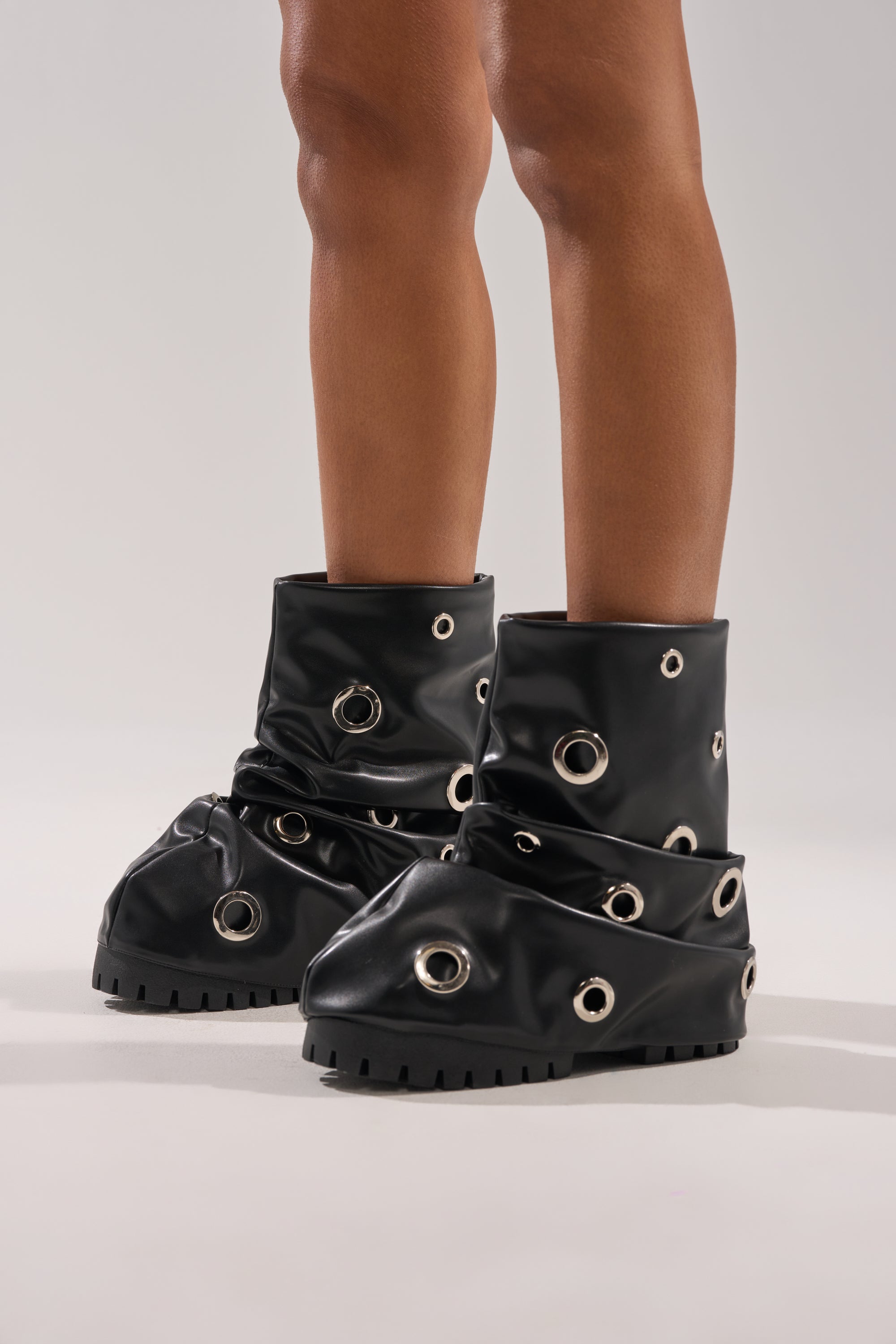 Only the lower legs and feet are visible, showcasing the AKIRA GOLD FABLE BLACK GROMMET EMBELLISHED FLAT BOOTIE with thick soles and large silver grommet holes, set against a light gray background.