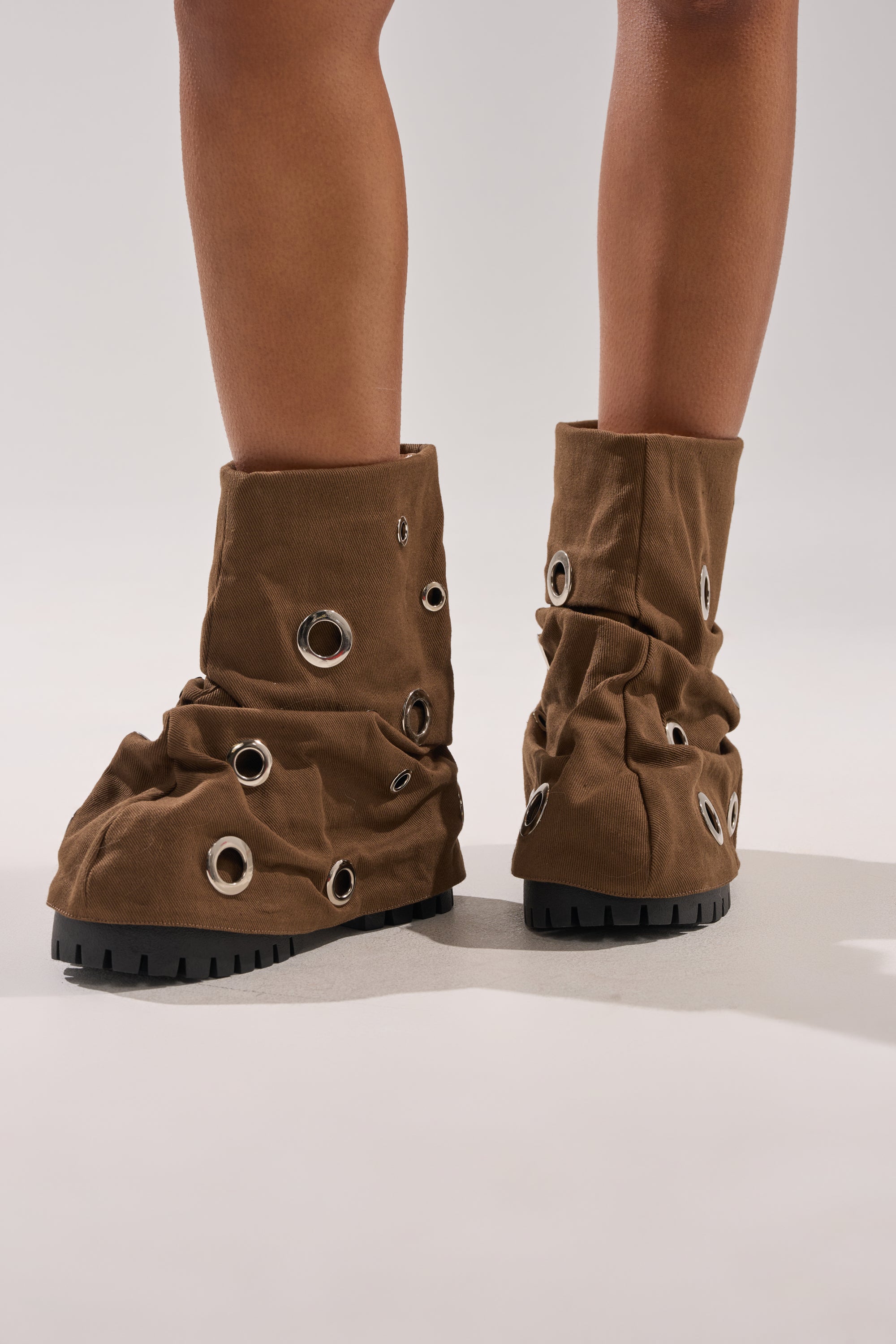 Someone stands on a neutral background wearing the AKIRA GOLD FABLE BROWN GROMMET EMBELLISHED FLAT BOOTIE—brown, slouchy booties with chunky black lug soles and large silver grommets across the fabric. 0