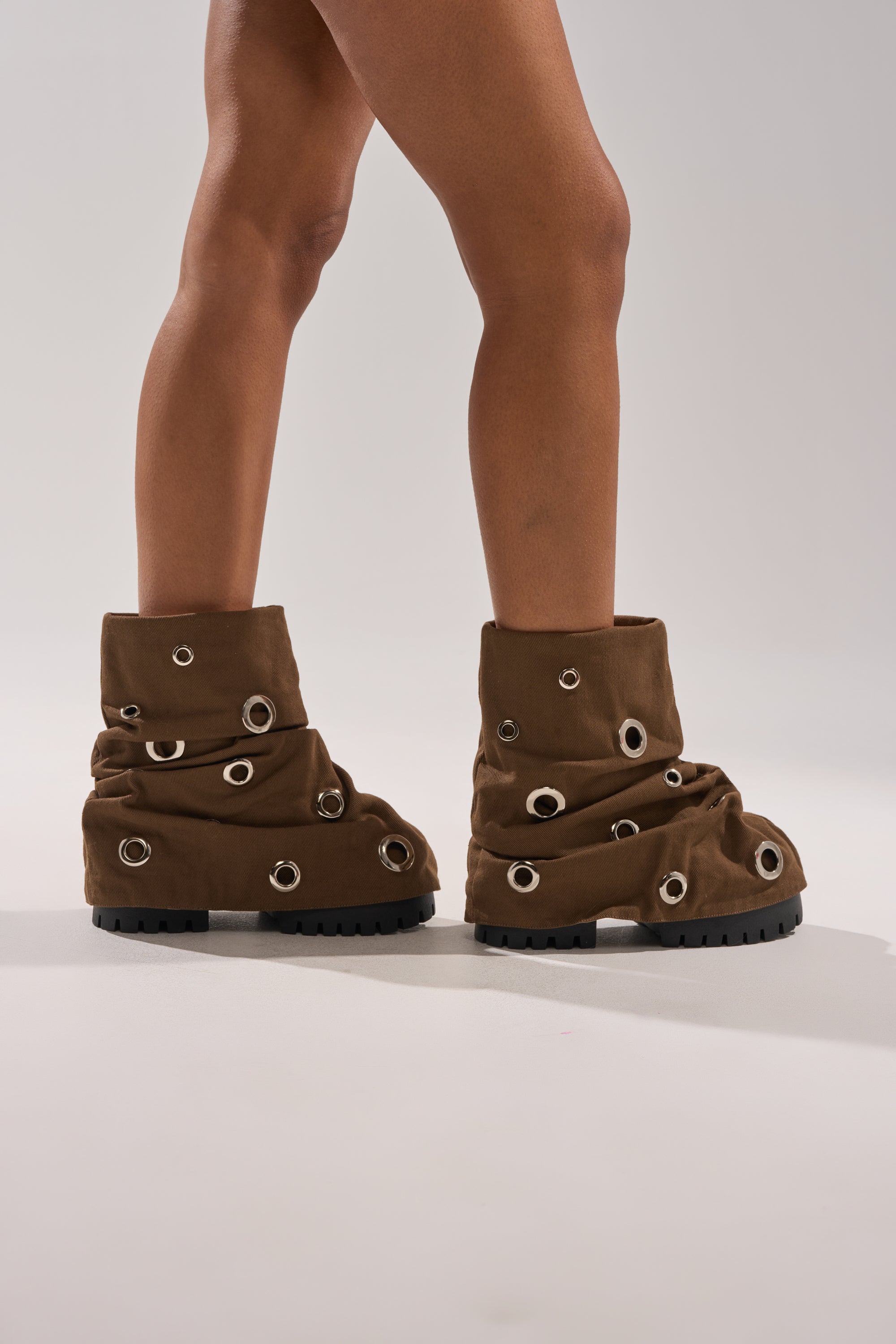 A person wearing AKIRA GOLD FABLE Brown Grommet Embellished Flat Booties stands against a plain light background. 1