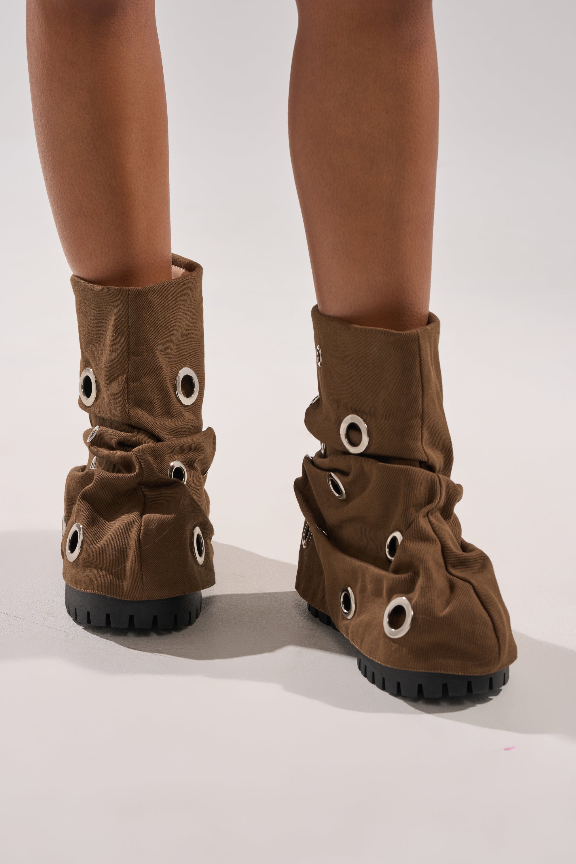 A person wearing AKIRA GOLD FABLE BROWN GROMMET EMBELLISHED FLAT BOOTIE with large silver grommets and chunky black soles stands on a light-colored surface. 2