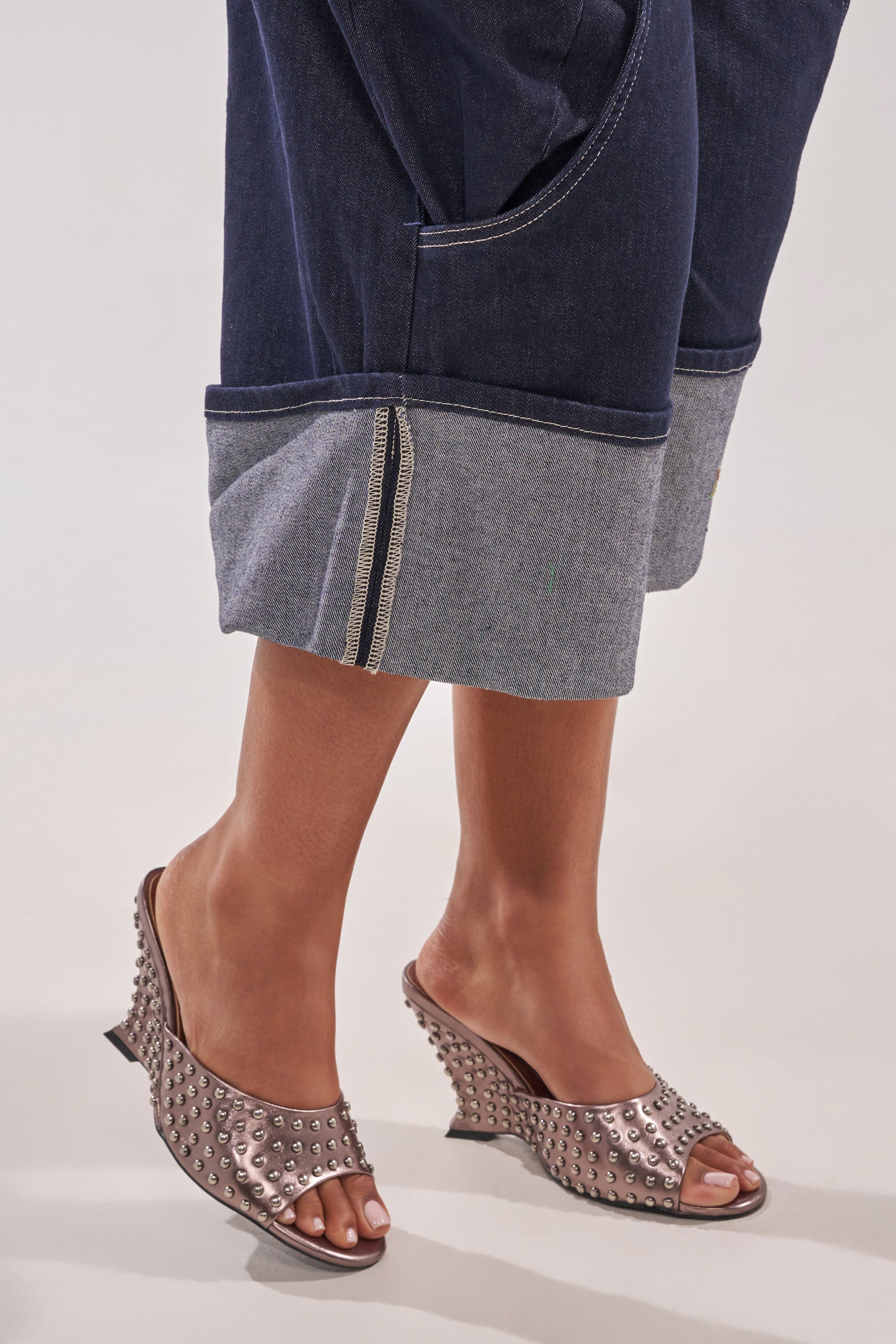 A person in cuffed dark denim pants with visible stitching wears AZALEA WANG FABRON PEWTER EMBELLISHED WEDGE SANDALS, standing against a plain light background. 0
