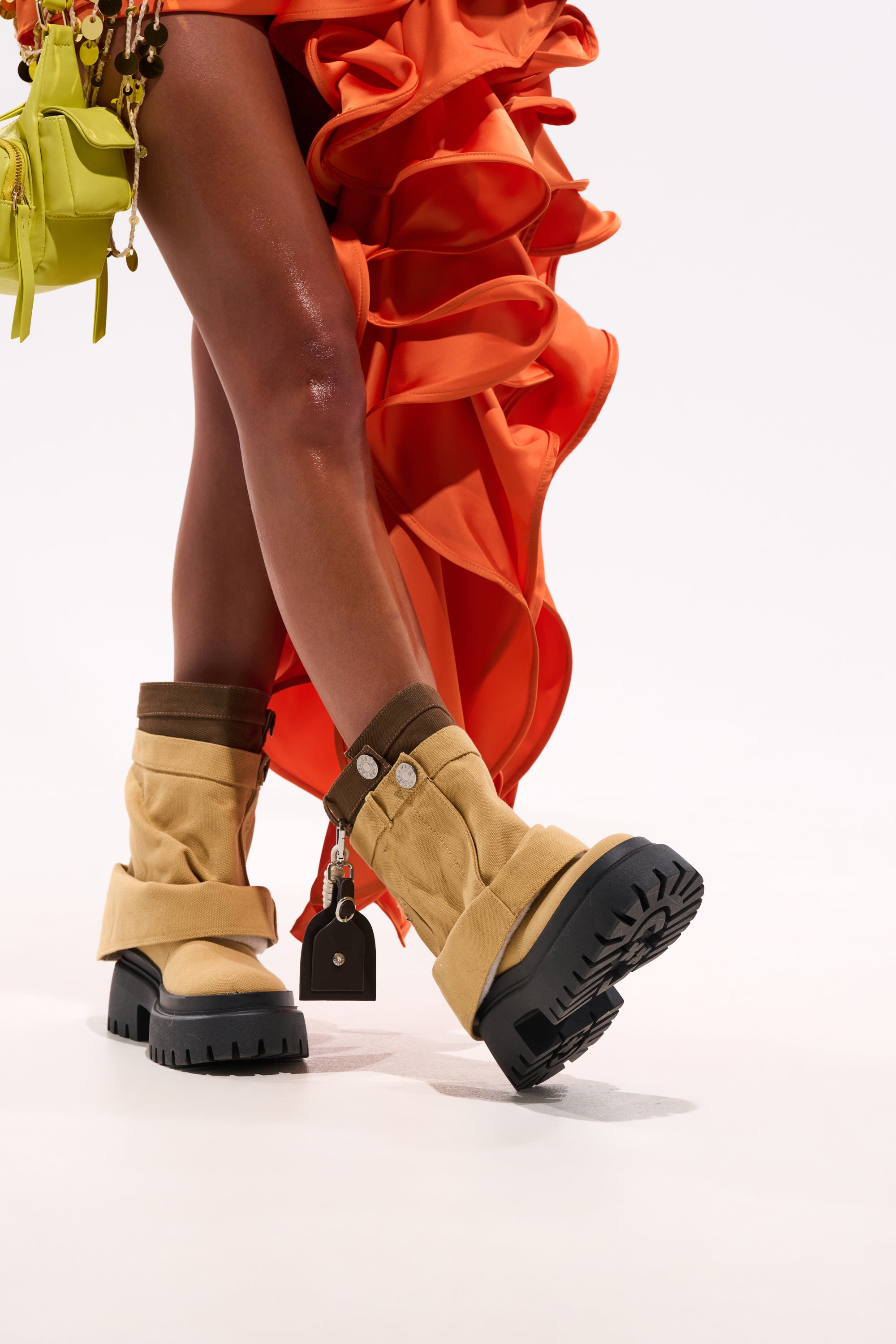 Wearing the AKIRA GOLD FAE TAN BOOTIE with chunky lug heels and thick black soles, a person pairs them with an orange ruffled skirt and a small yellow bag with decorative keychains, standing against a white background. 0