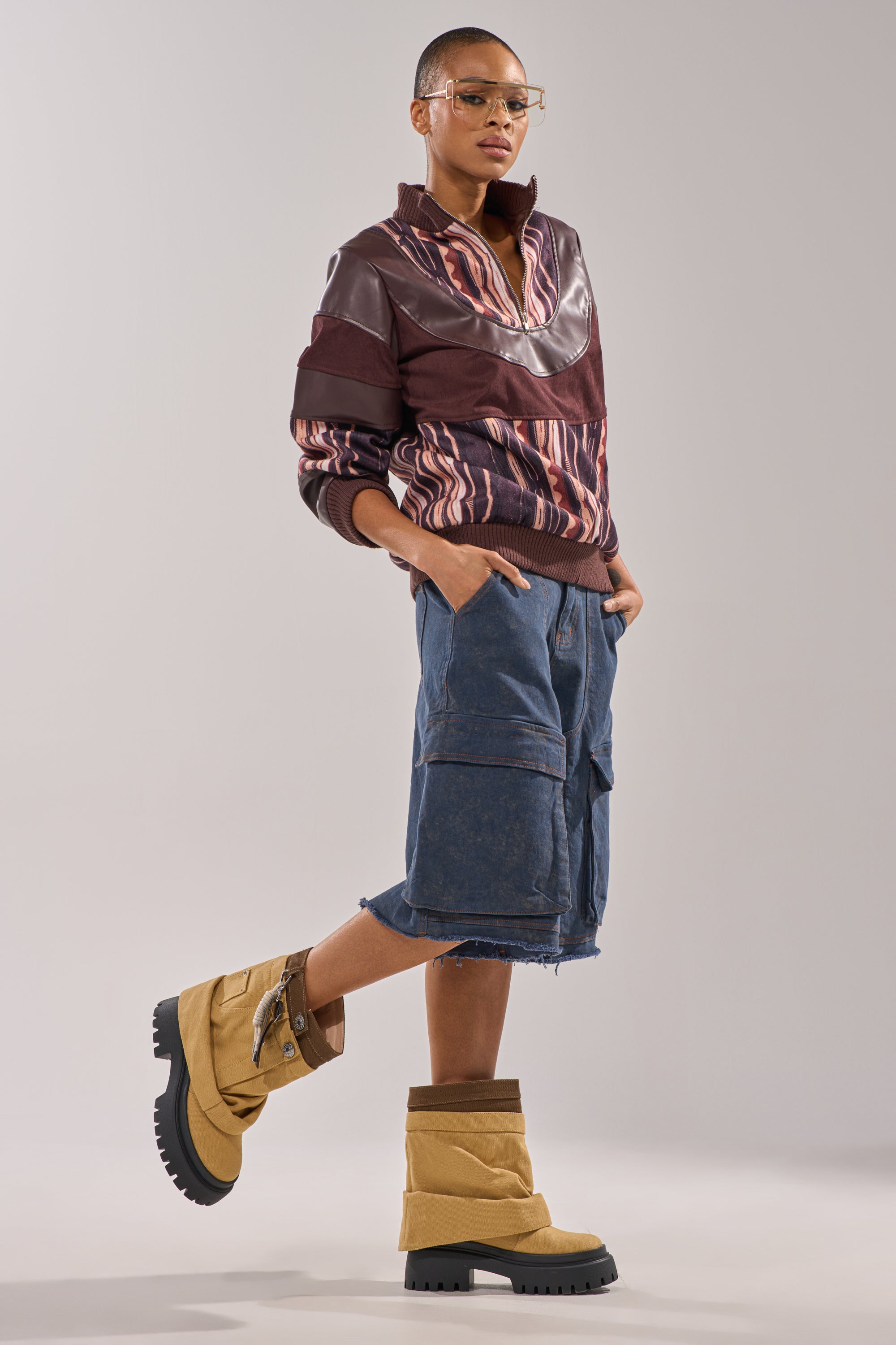Wearing glasses, a maroon patterned jacket, denim shorts, and AKIRA GOLD FAE TAN BOOTIE with chunky lug heel and platform sole, a short-haired person stands against a plain backdrop with one foot raised on tiptoe. 3