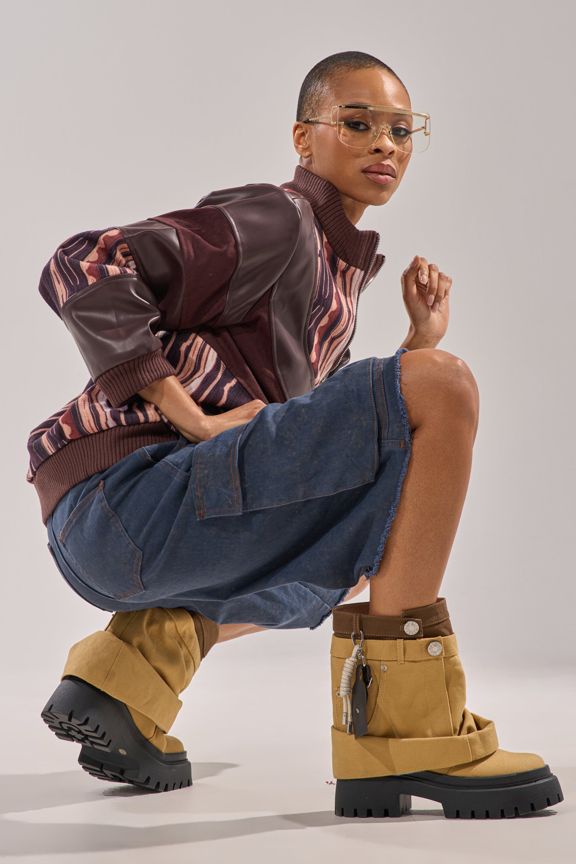 A person squats confidently in a patterned jacket, baggy denim shorts, and AKIRA GOLD FAE TAN BOOTIE with a chunky lug heel and oversized clear glasses, set against a plain studio background. 8
