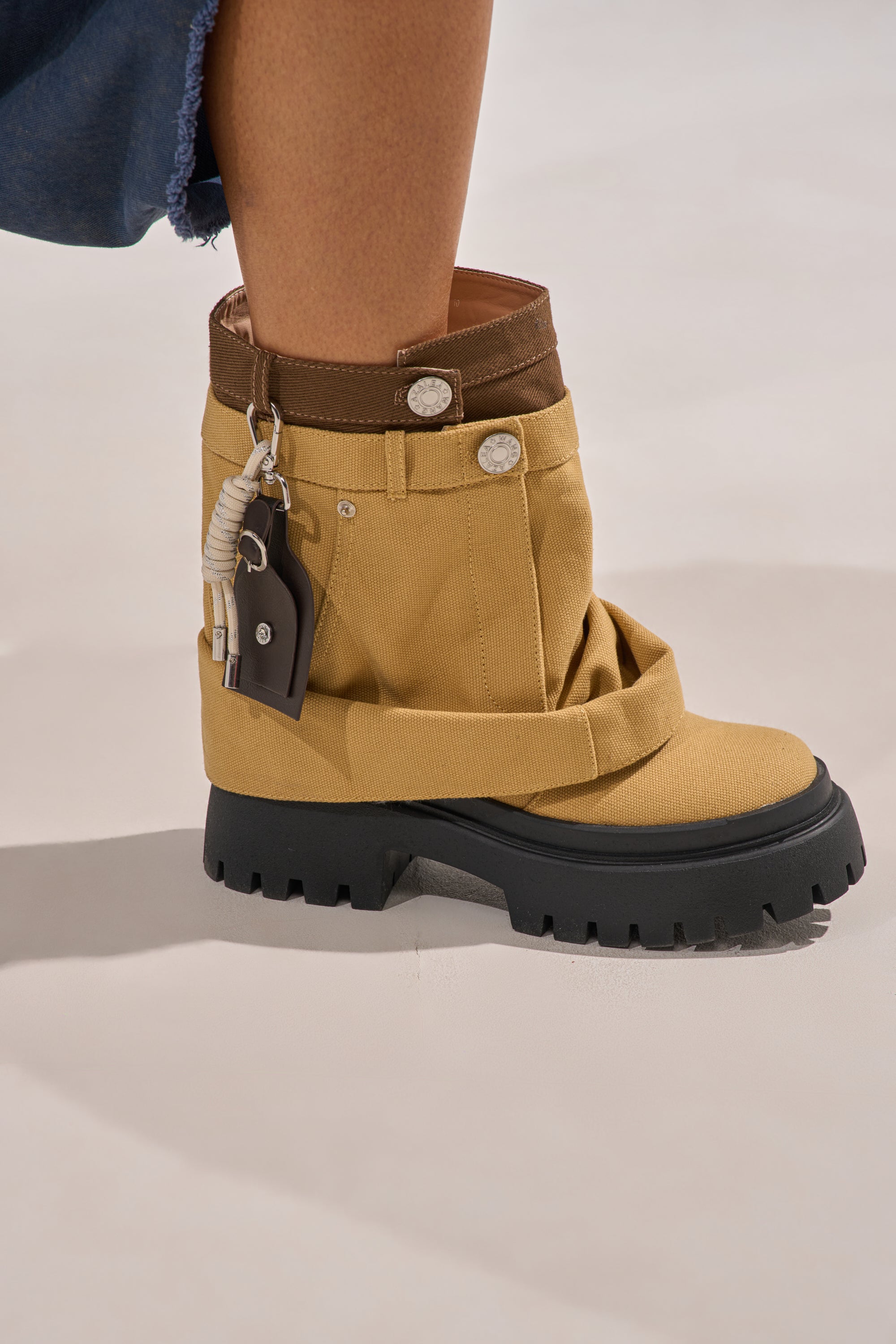 A close-up of the AKIRA GOLD FAE TAN BOOTIE, featuring tan denim, a chunky lug heel, thick black platform sole, decorative buttons, and keychain pouch, worn by someone in a blue outfit. 9