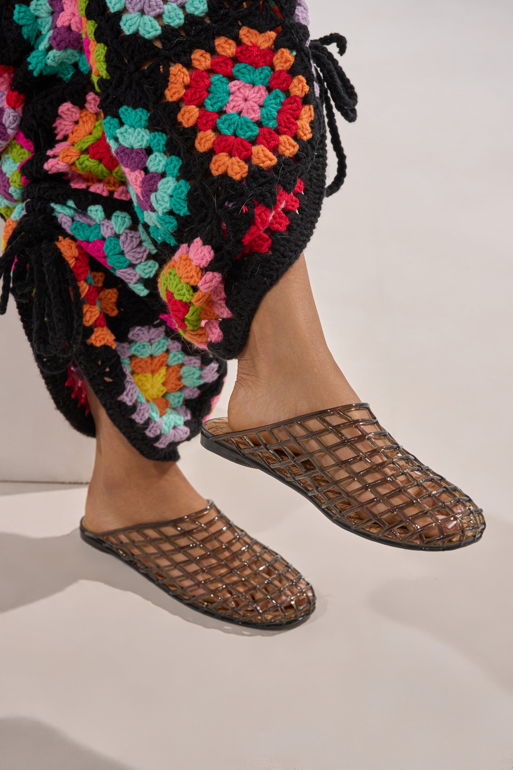 A person wearing a colorful crocheted garment and the AKIRA FAIROZE BLACK JELLY MULE sandals, their feet slightly off the ground against a plain background. 0