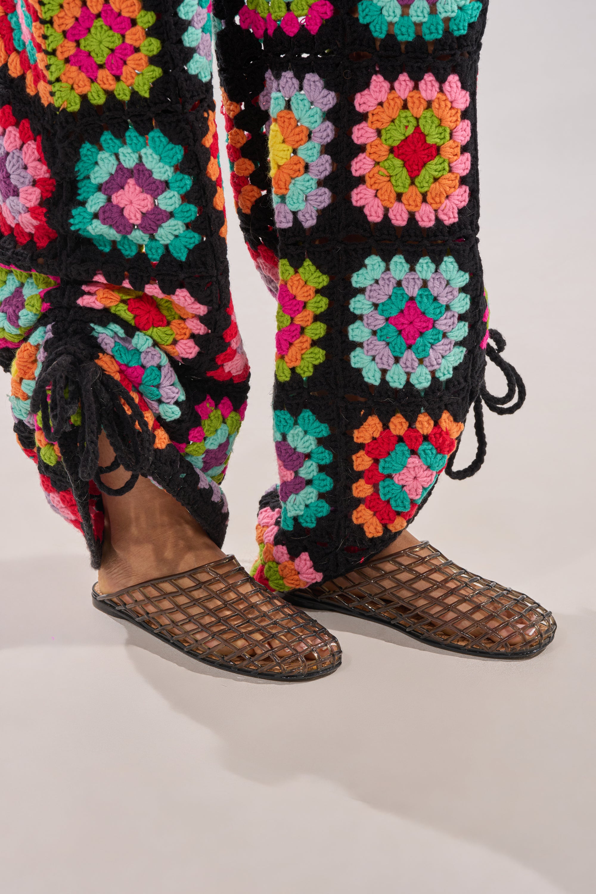 Close-up of a person wearing colorful crocheted pants with a granny square pattern and black drawstrings at the ankles, paired with the AKIRA FAIROZE BLACK JELLY MULE. The background is plain and light-colored. 1