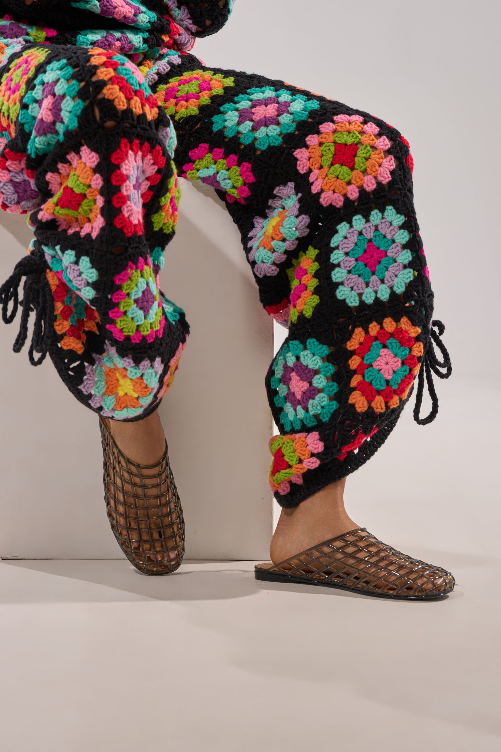 A person sits on a white block, showing only their lower half in colorful square-pattern crochet pants and stylish AKIRA FAIROZE BLACK JELLY MULES—perfect summer footwear. 2