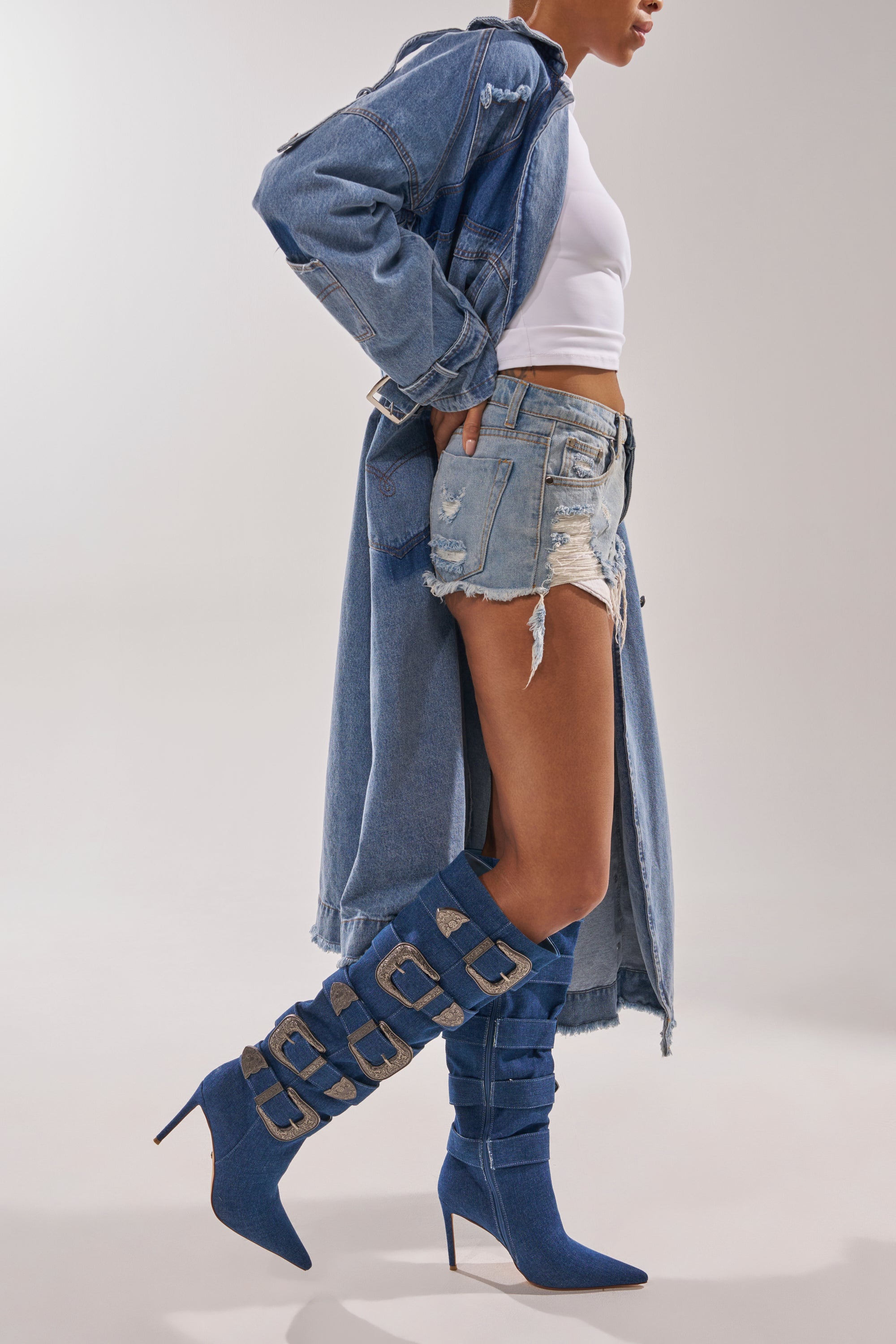 A person wears a long denim jacket, white crop top, distressed shorts, and AKIRA GOLD FALLON DENIM BUCKLE EMBELLISHED STILETTO BOOTS with bold buckles, standing against a plain light background. 1
