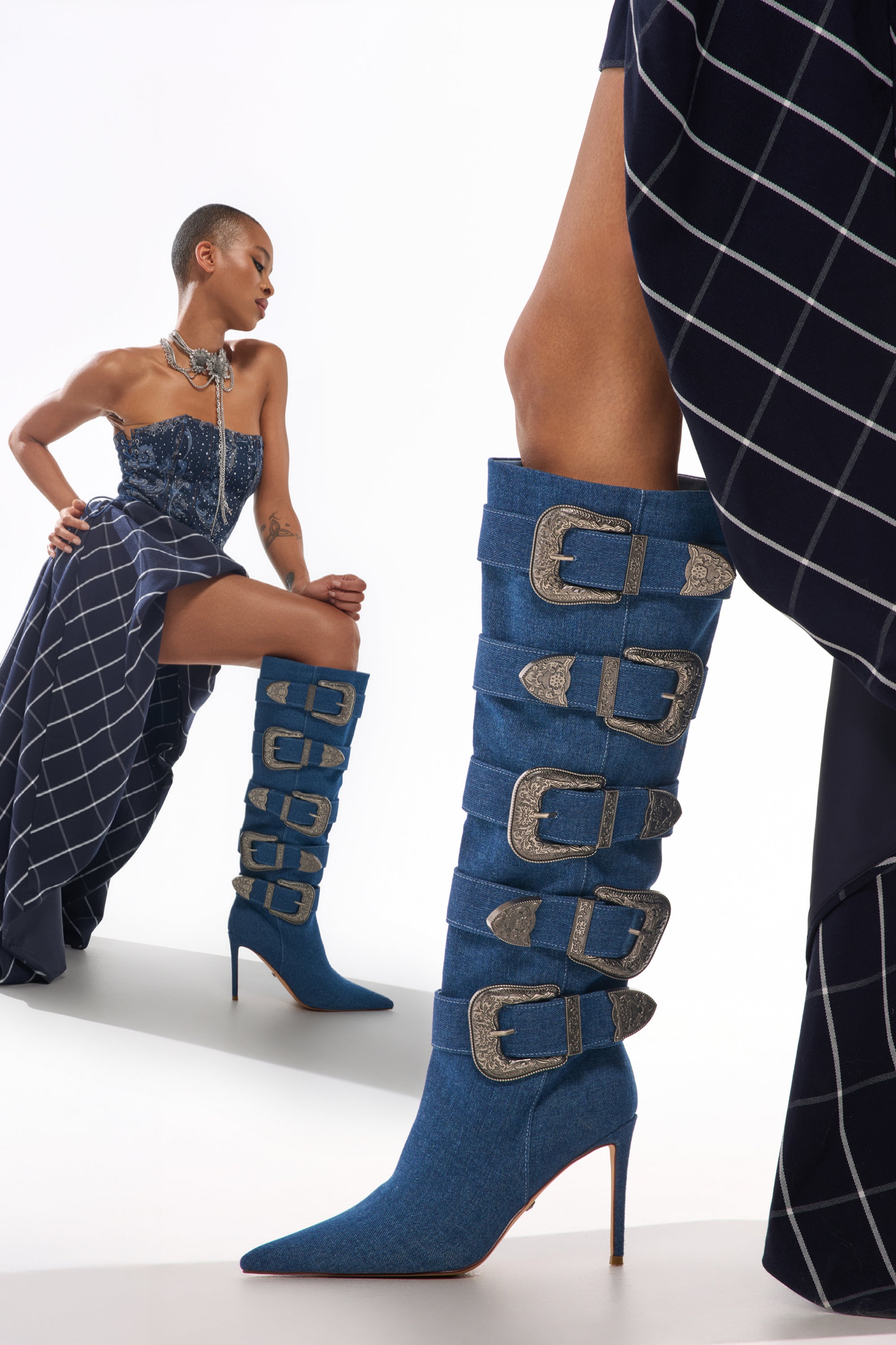 A model poses in a blue grid-patterned strapless dress, showcasing the AKIRA GOLD FALLON DENIM BUCKLE EMBELLISHED STILETTO BOOT—tall, western-inspired with high heels and bold silver buckles. 0