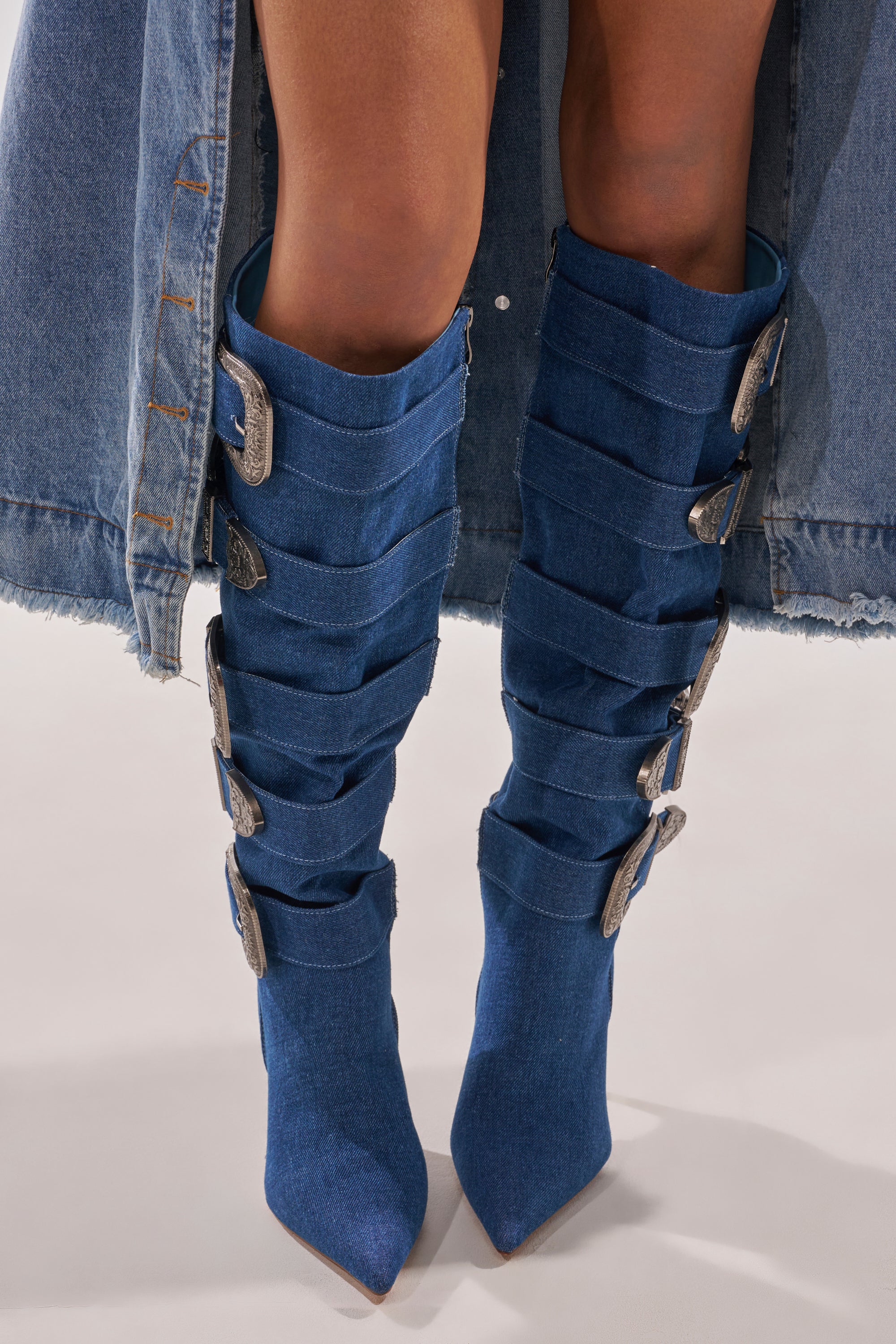 A person wears the AKIRA GOLD FALLON DENIM BUCKLE EMBELLISHED STILETTO BOOT—knee-high, pointed toe, with wide straps and silver buckles—paired with a frayed denim skirt for a modern cowgirl couture look. 2