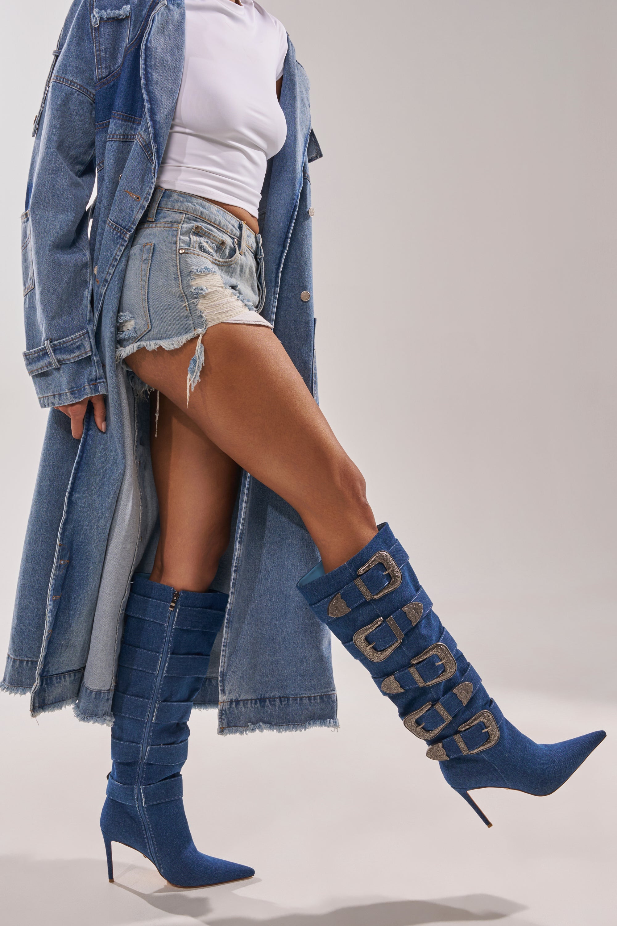 A person wears a long denim coat, distressed shorts, a white crop top, and bold knee-high AKIRA GOLD FALLON DENIM BUCKLE EMBELLISHED STILETTO BOOTS, channeling cowgirl couture against a plain background. 3