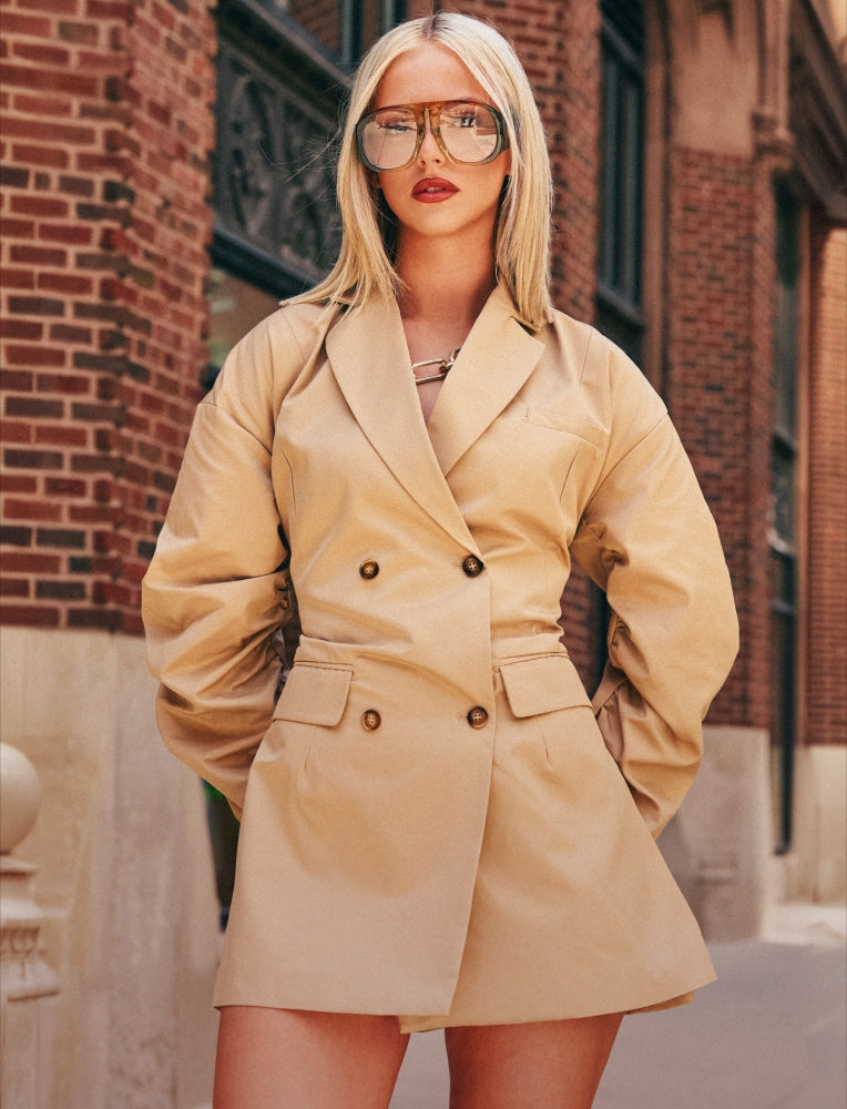 A blonde woman wearing a beige double-breasted blazer-style mini dress with oversized sunglasses, standing outdoors in front of a brick building.