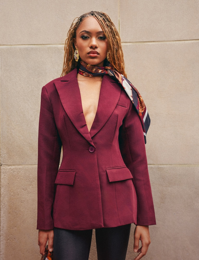 A woman wearing a tailored burgundy blazer with a deep V-neckline, paired with a patterned scarf and dark pants, standing against a beige stone wall.