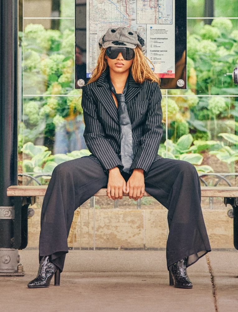 A model sits on a bench at a transit station wearing a black pinstripe blazer, matching wide-leg trousers, a gray tie, a gray beanie, black sunglasses, and black boots.