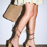A person in a light yellow dress and AZALEA WANG FANTASA BROWN HALLOW PLATFORM WEDGE STRAPPY SANDALS with ankle ties and chain details, holding a woven straw tote bag, stands against a plain white background.