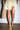 A woman wears the AZALEA WANG FANTASA BROWN HALLOW PLATFORM WEDGE STRAPPY SANDAL with a light yellow dress and carries a woven straw bag, standing on polished concrete against a plain white wall.