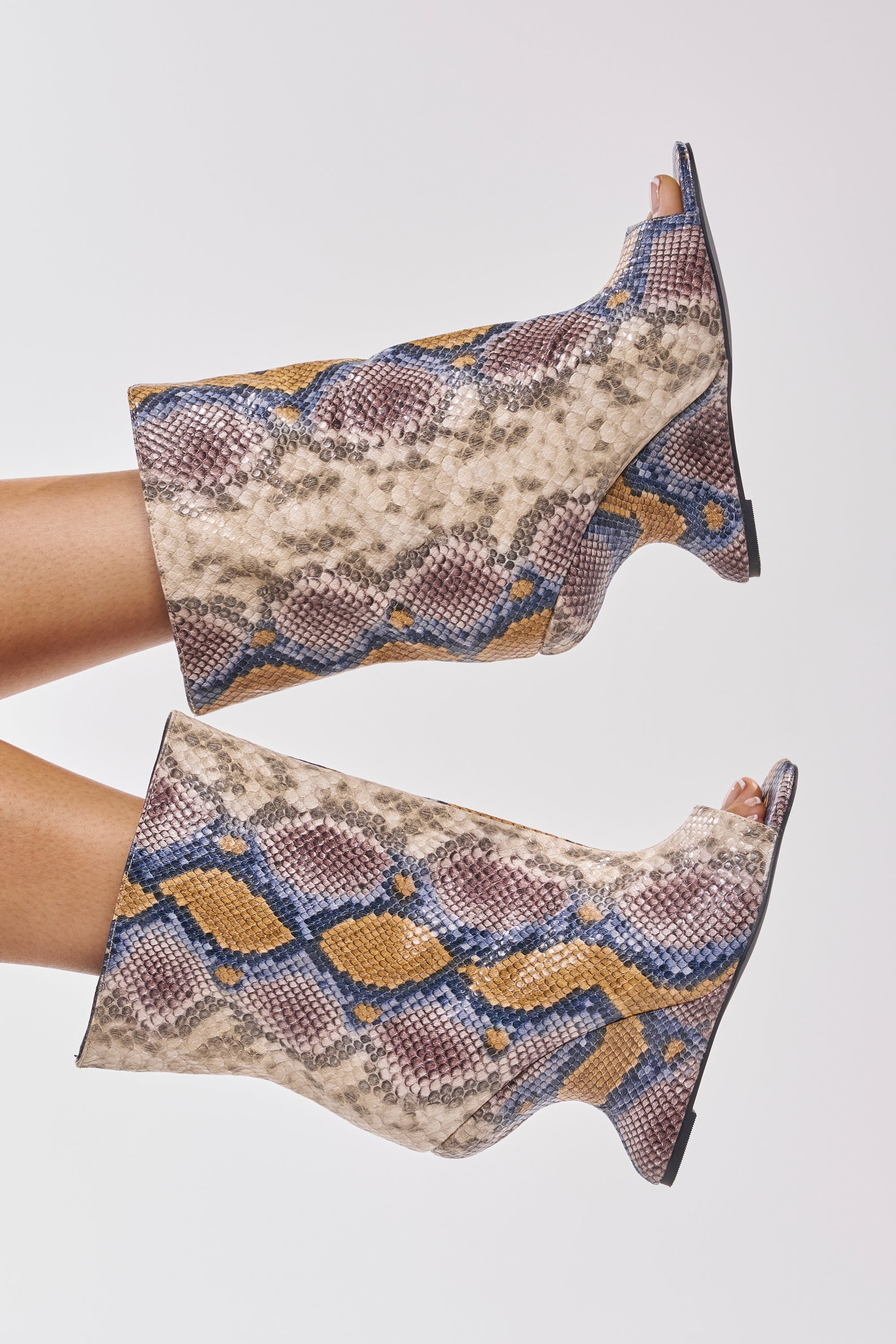 A pair of legs models the AZALEA WANG FAOI SNAKE MULTI OPEN TOE WEDGE BOOTIE, featuring a snake print in tan, yellow, blue, and purple faux leather with an inverted wedge heel, shown against a plain white background. 0